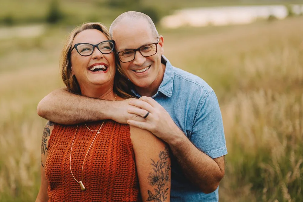 A middle-aged woman with glasses and tattoos smiling and hugging a man with glasses and a blue shirt outdoors in a field.