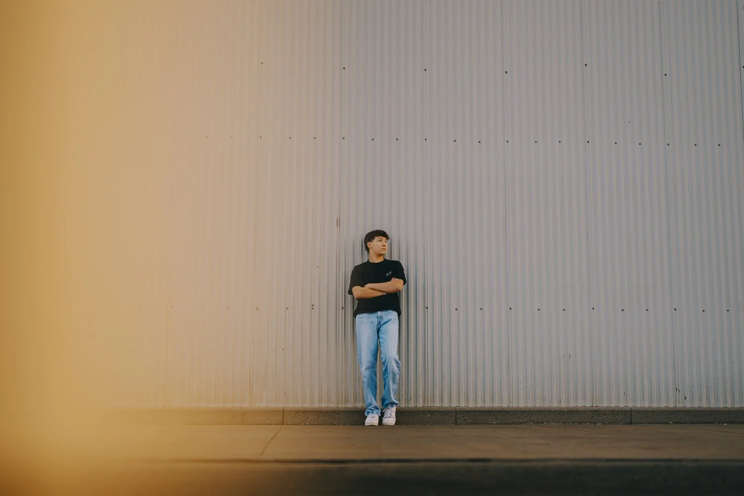 A person standing with arms crossed against a large, corrugated metal wall, wearing a black t-shirt, light blue jeans, and white sneakers.