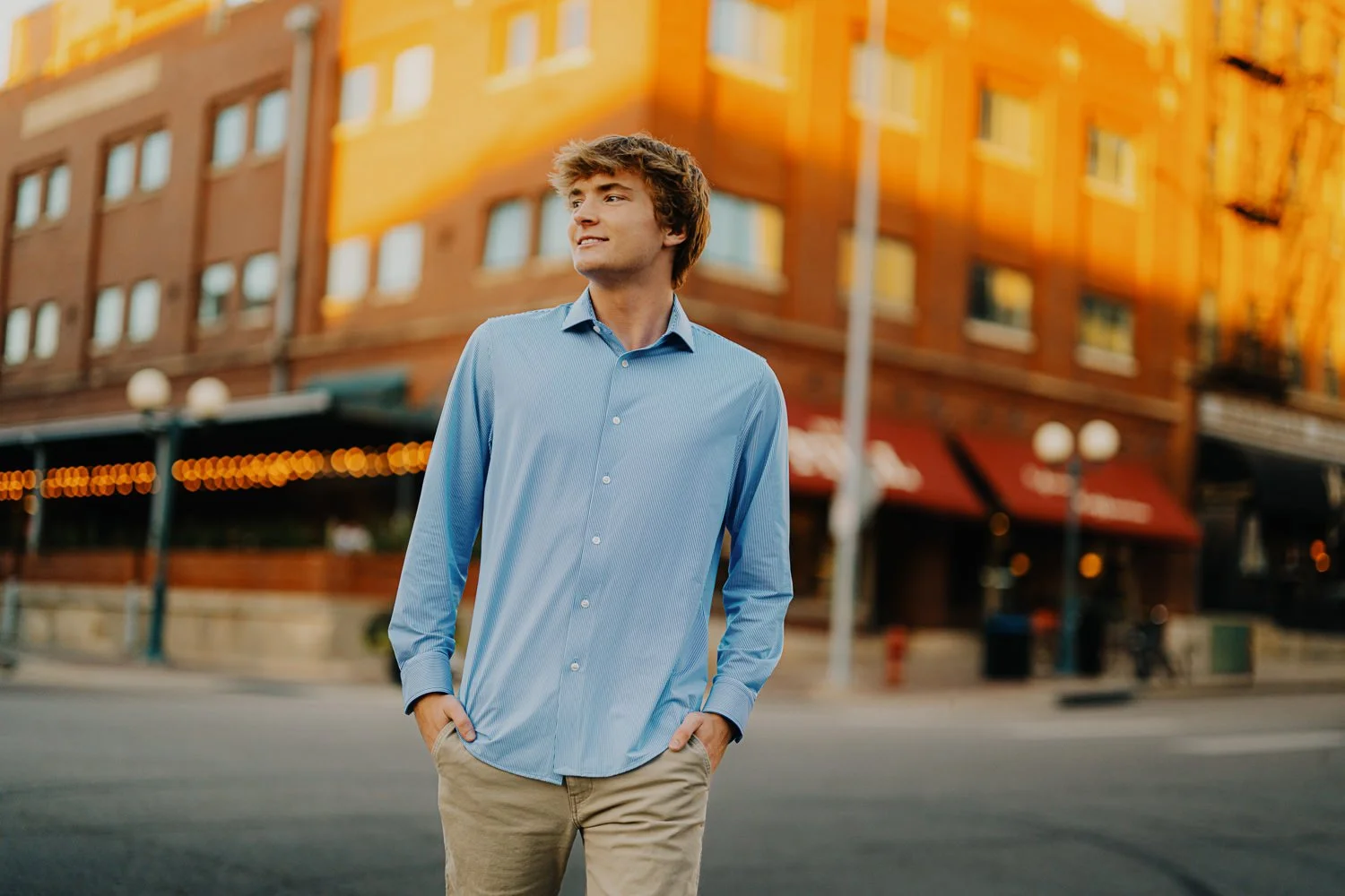 A young man in a light blue button-down shirt and tan pants stands on a city street in front of a brick building with warm lighting during sunset.