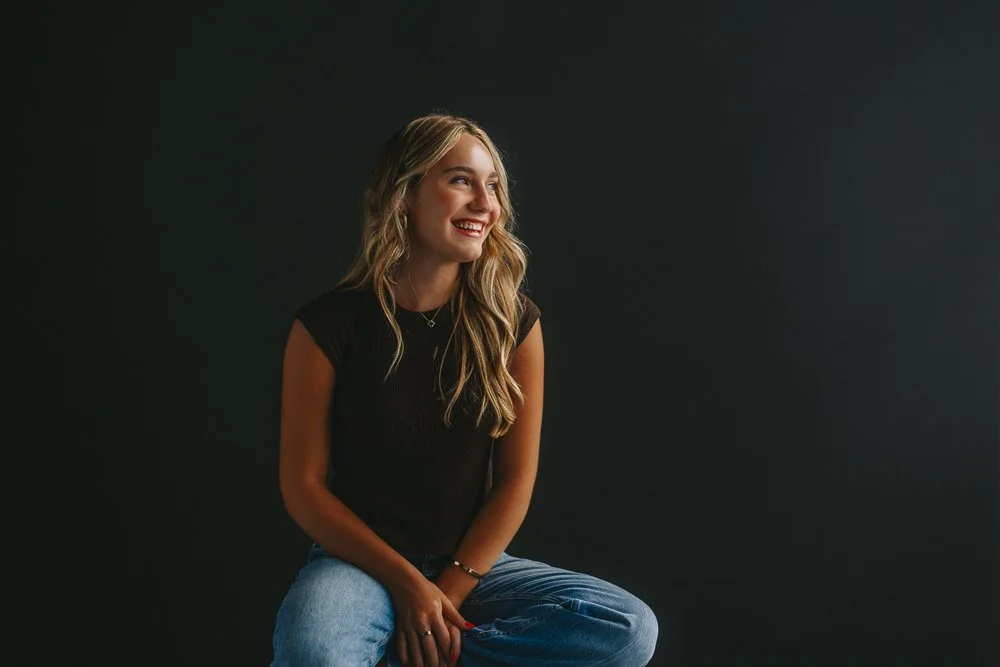 Smiling woman with long blonde hair wearing a black top and blue jeans, sitting against a dark background.