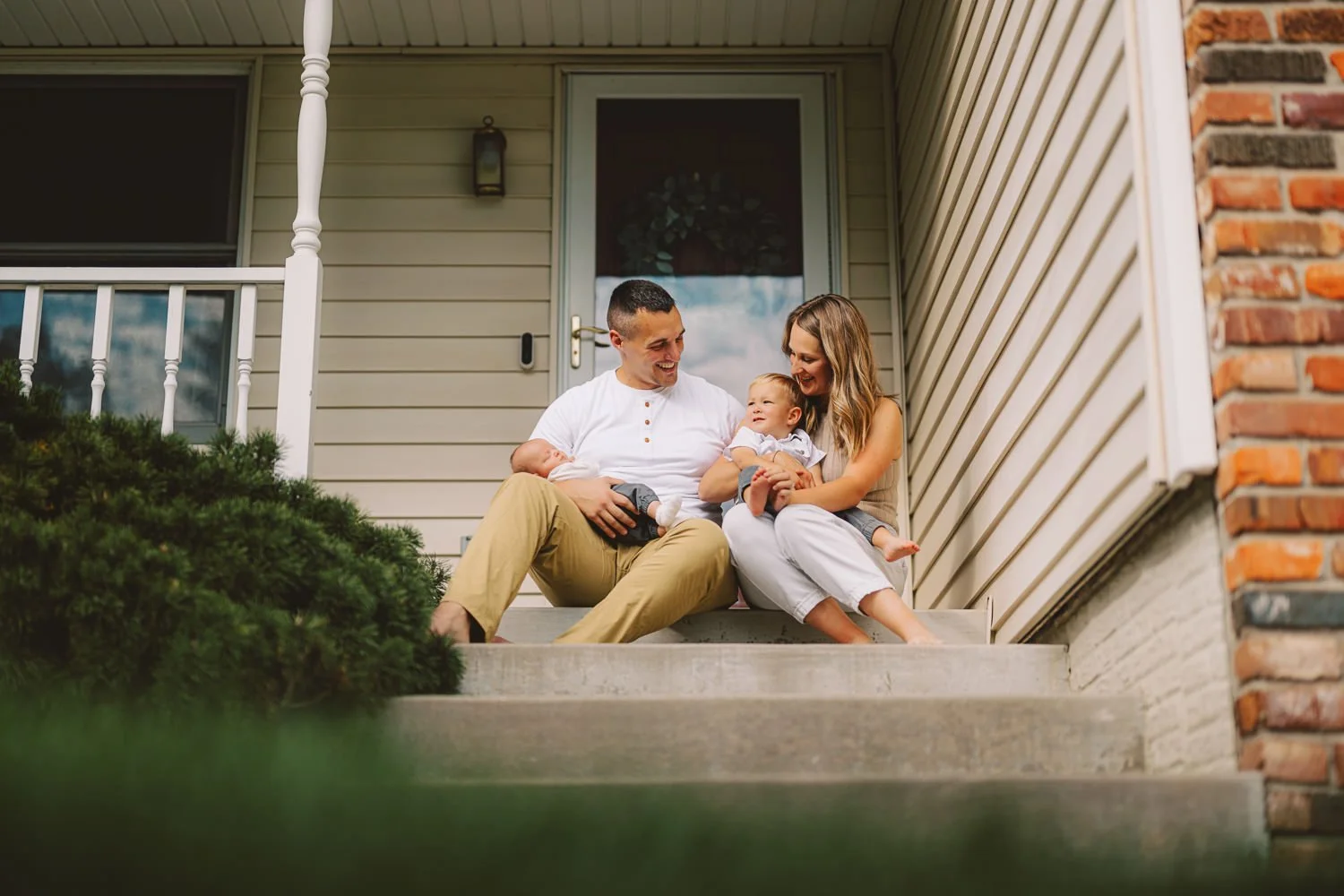 A happy family sitting on their porch steps, with two adults and two young children, smiling and interacting.