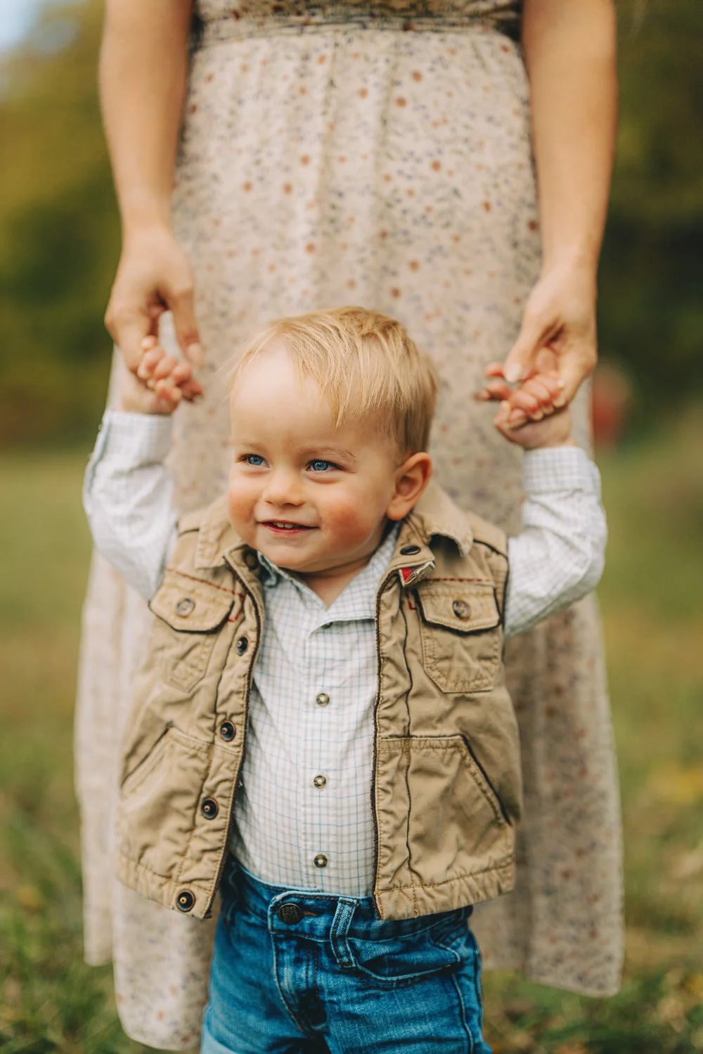 A young boy with blonde hair and blue eyes, smiling, holding hands with an adult woman outdoors. The woman is wearing a floral dress and the boy is dressed in a checked shirt, tan vest, and blue jeans.