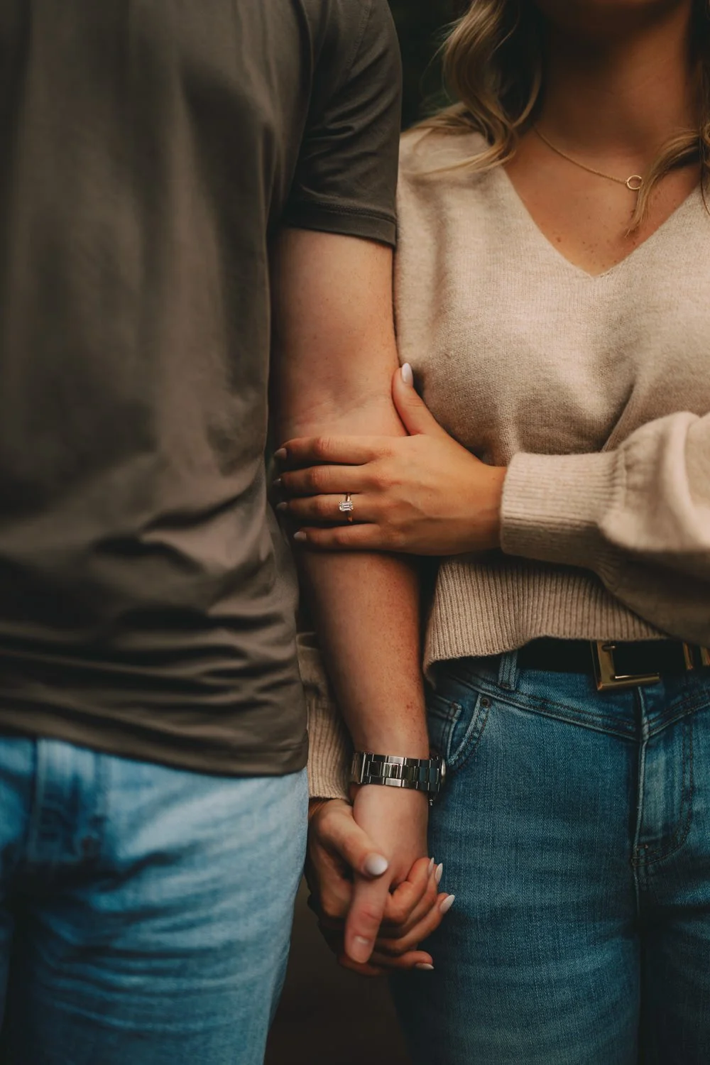 A close-up of a couple holding hands with the woman's hand showing a ring on her finger. The woman is wearing a beige sweater, and the man is wearing a gray t-shirt and blue jeans.