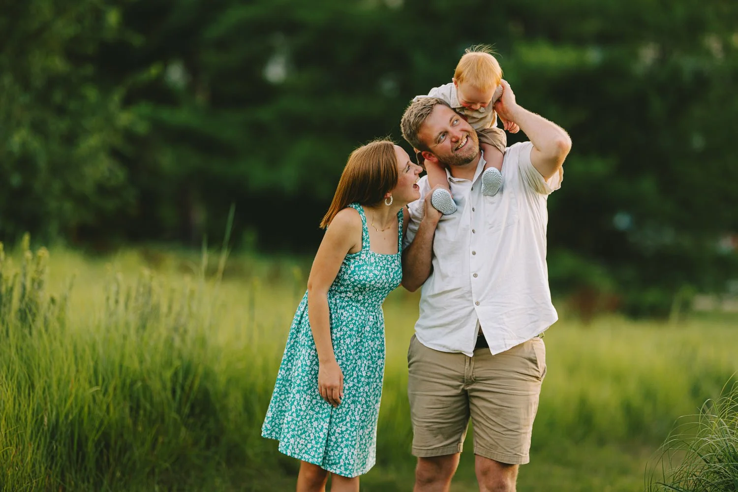 A family of three enjoying time outdoors in a grassy field with trees in the background. The father is carrying a young child on his shoulders, the mother is standing close and smiling at them.