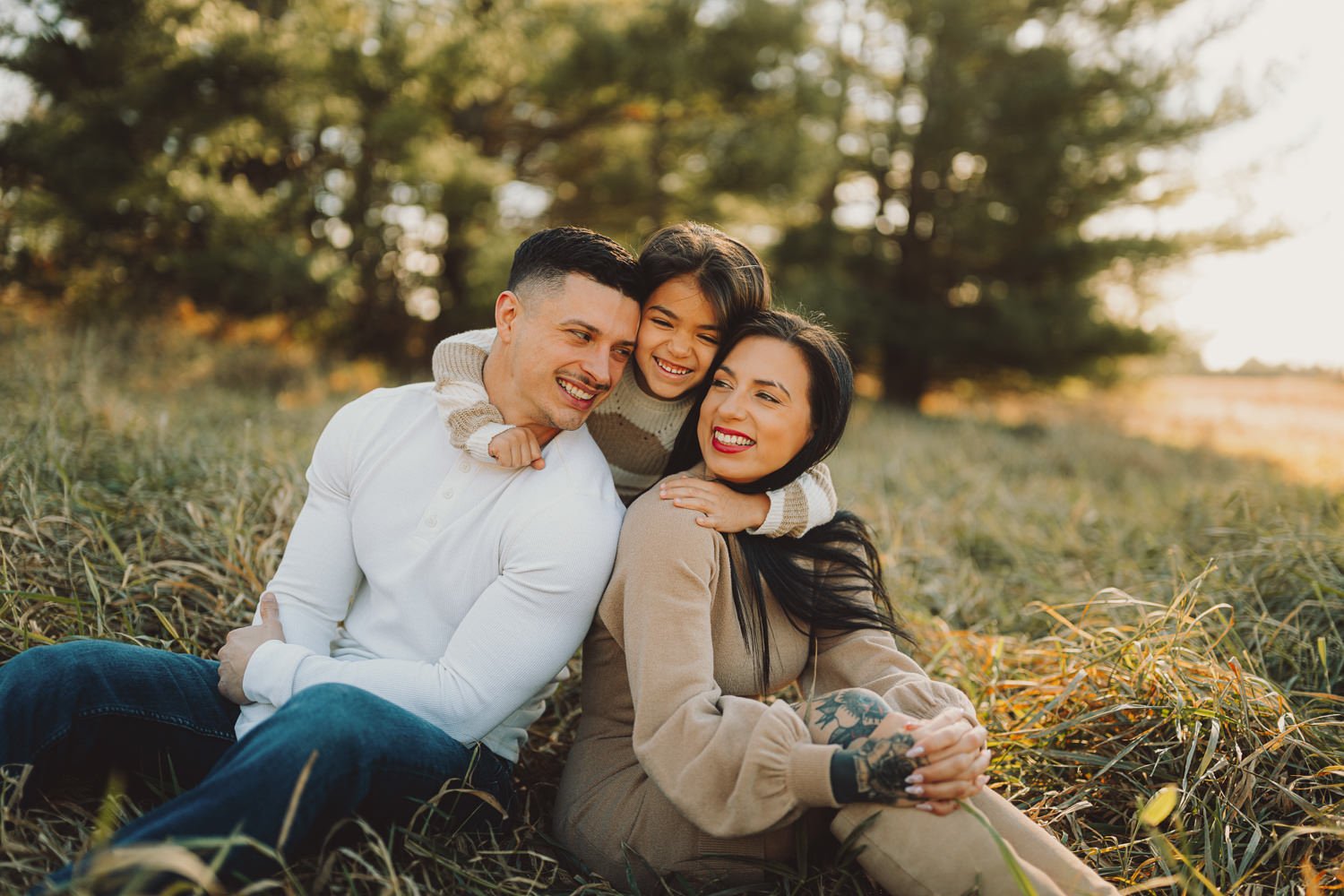 A happy family of three sitting on grass in a field during sunset, with the child leaning on the father and smiling, the mother smiling at the camera, all sharing a joyful moment.