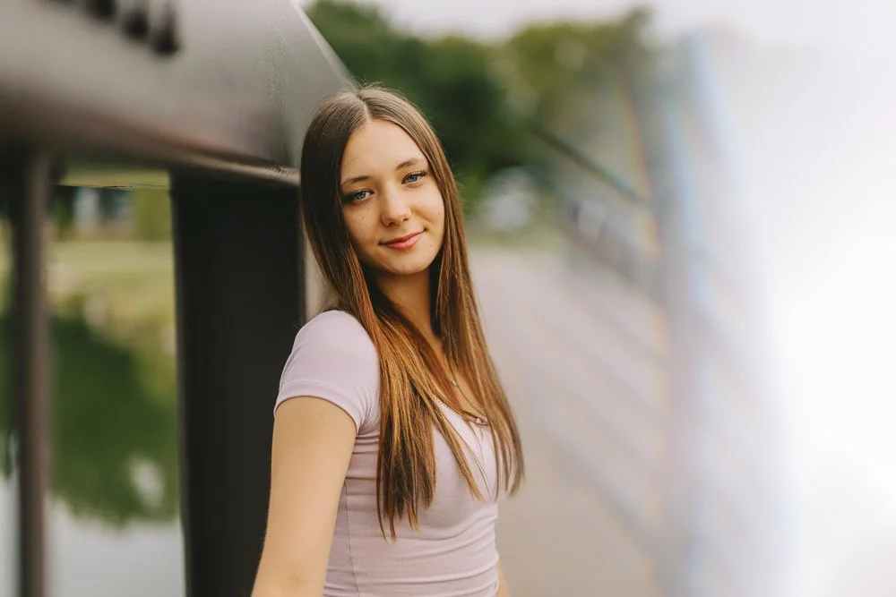A young woman with long brown hair leaning against a black railing outdoors near a body of water, smiling softly.