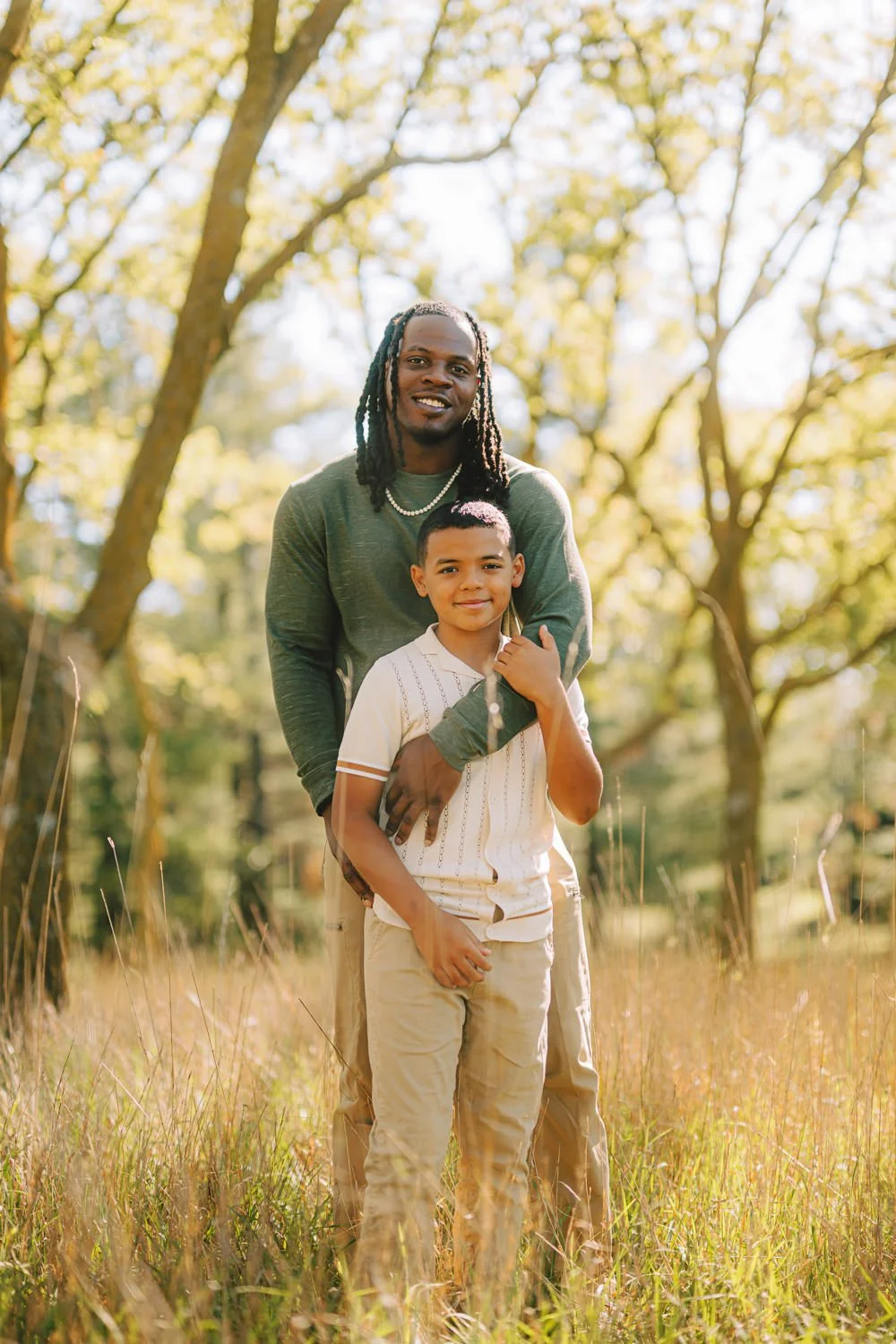 A man with dreadlocks and a young boy stand outdoors in a grassy field with trees and sunlight in the background, smiling at the camera.
