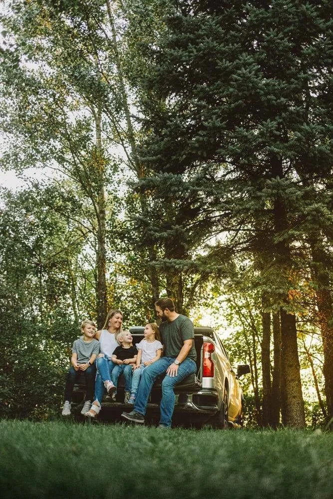 Family sitting on the back of a pickup truck in a wooded area during sunset, smiling and talking.
