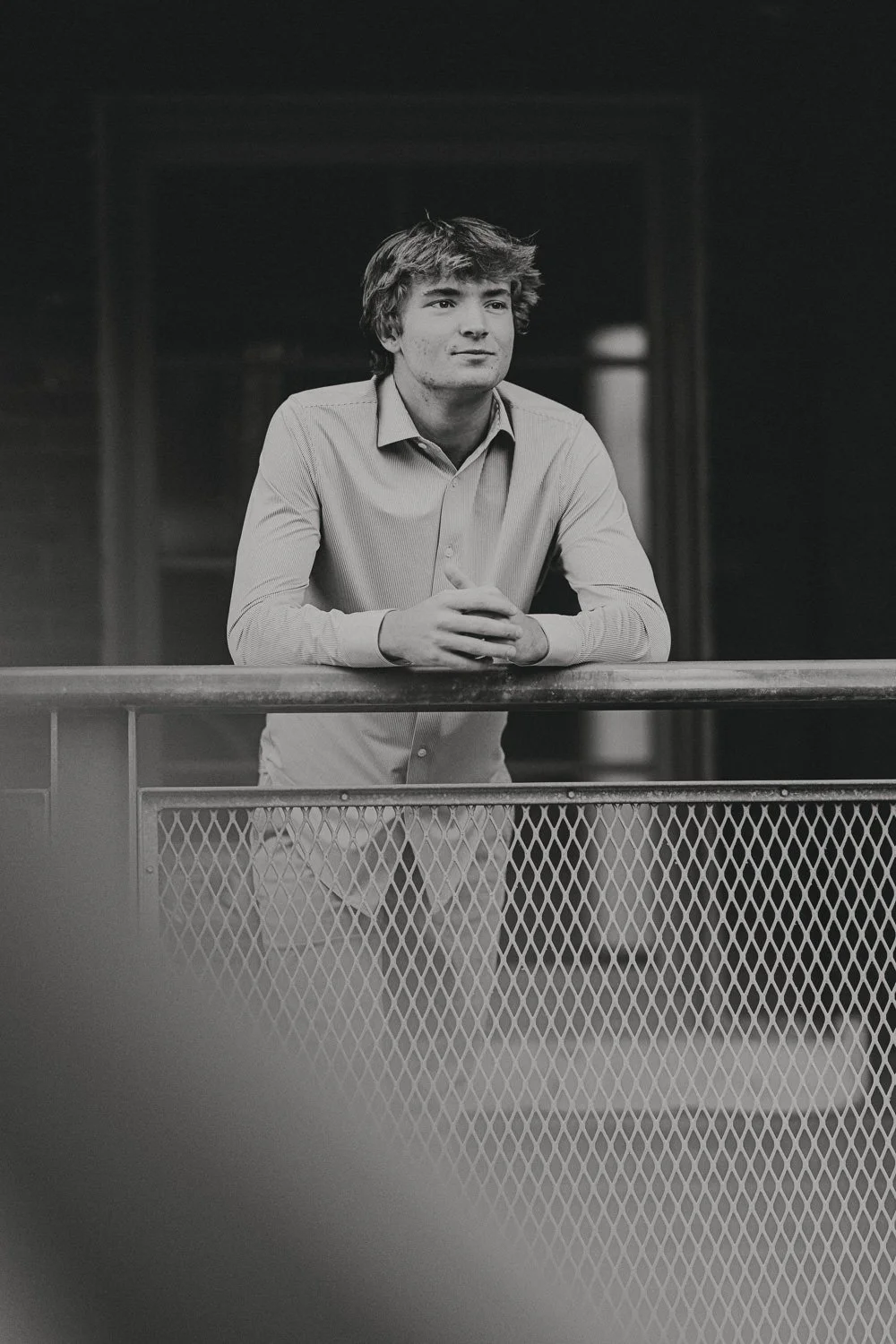 A young man with wavy hair wearing a button-up shirt, leaning on and holding his hands together on a railing, looking thoughtfully into the distance in a black-and-white photo.