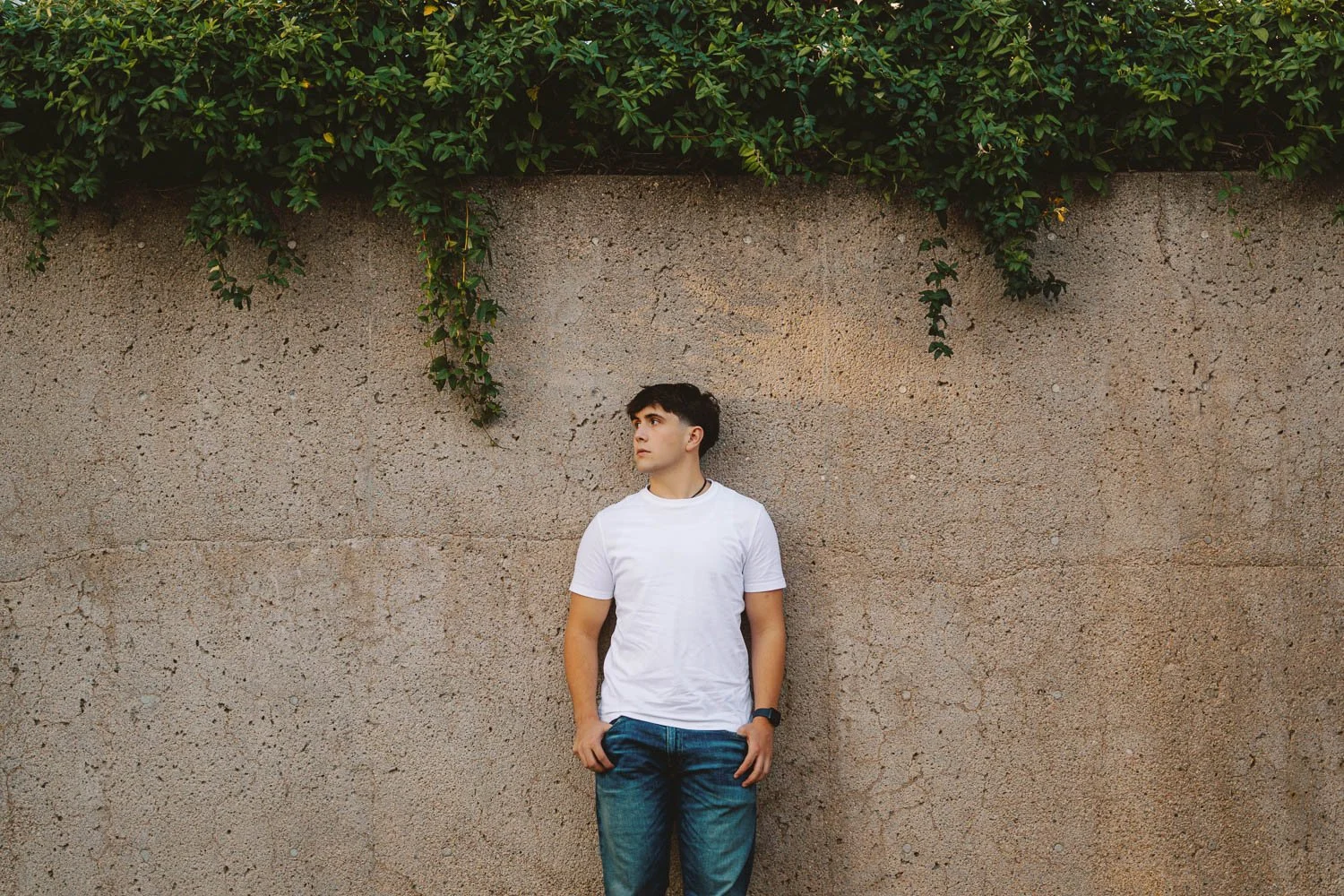 Young man wearing a white t-shirt and blue jeans standing against a textured beige wall with green foliage hanging above.