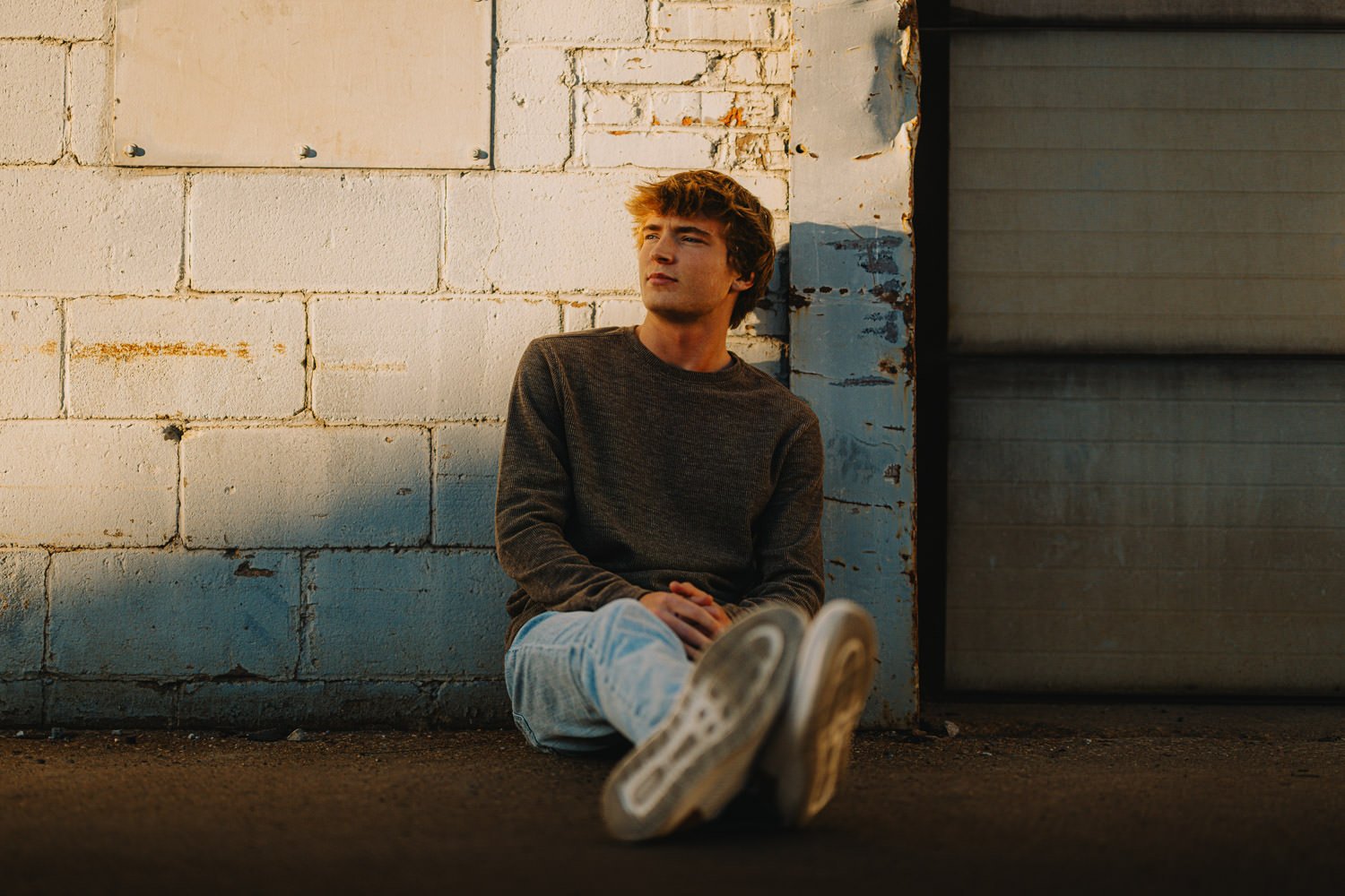 Young man sitting on the ground against a brick wall with a contemplative expression.