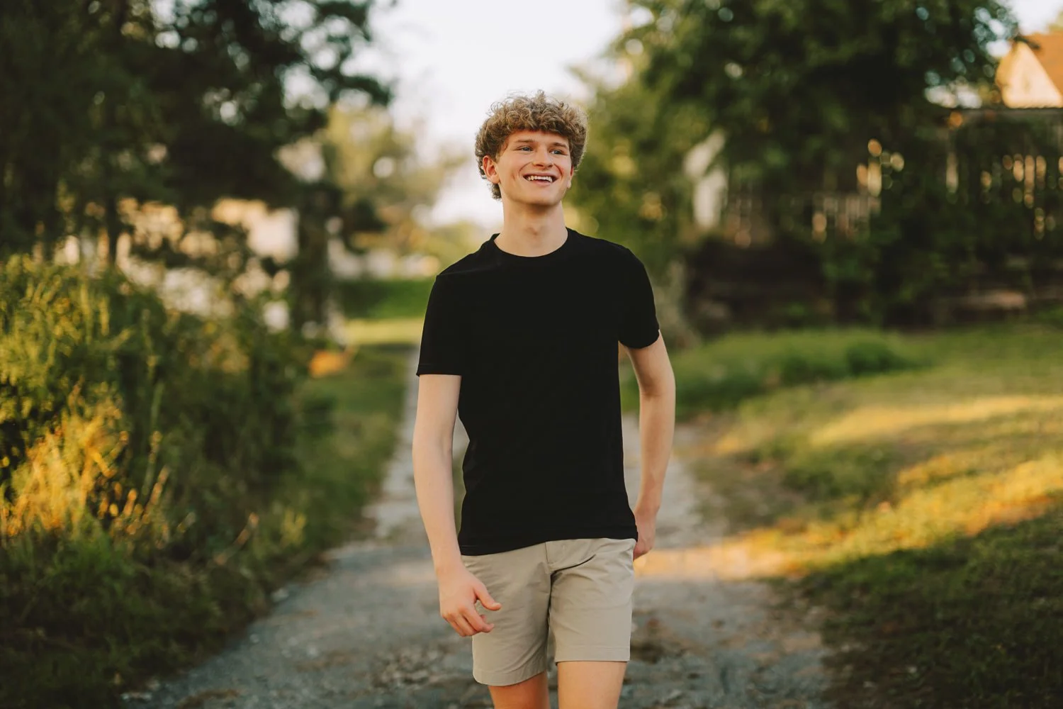 A young man with curly hair wearing a black t-shirt and beige shorts walking outdoors on a dirt path surrounded by greenery on a sunny day.