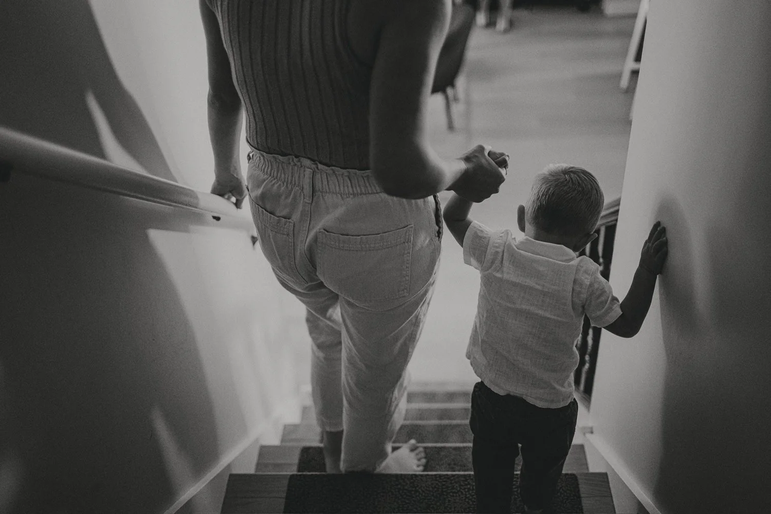 A woman and a young boy walk down a staircase together, holding hands. The image is in black and white.