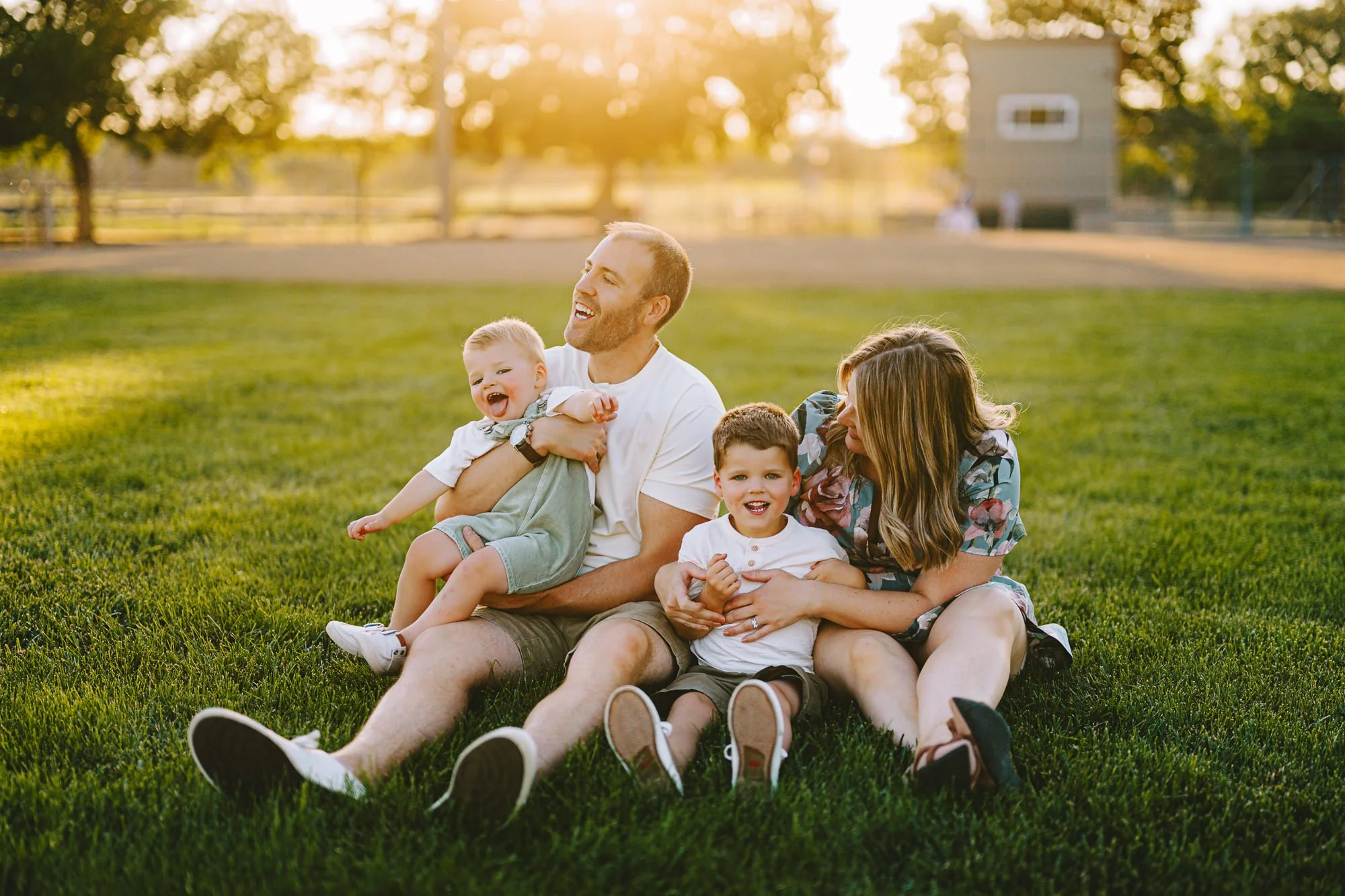 A happy family of four sitting on grass at a park during sunset, smiling and playing together.