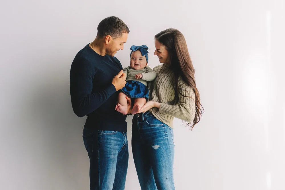 A happy family consisting of a man, woman, and baby girl standing together against a plain white wall.