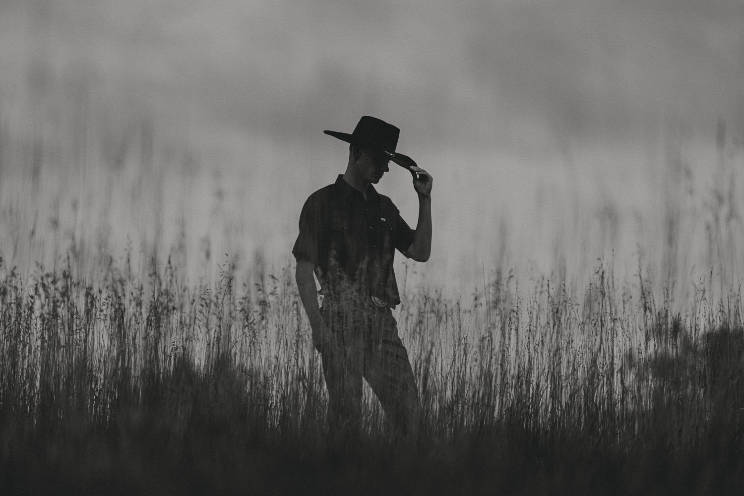 Silhouette of a man wearing a hat standing in a field of tall grass with a blurred background.