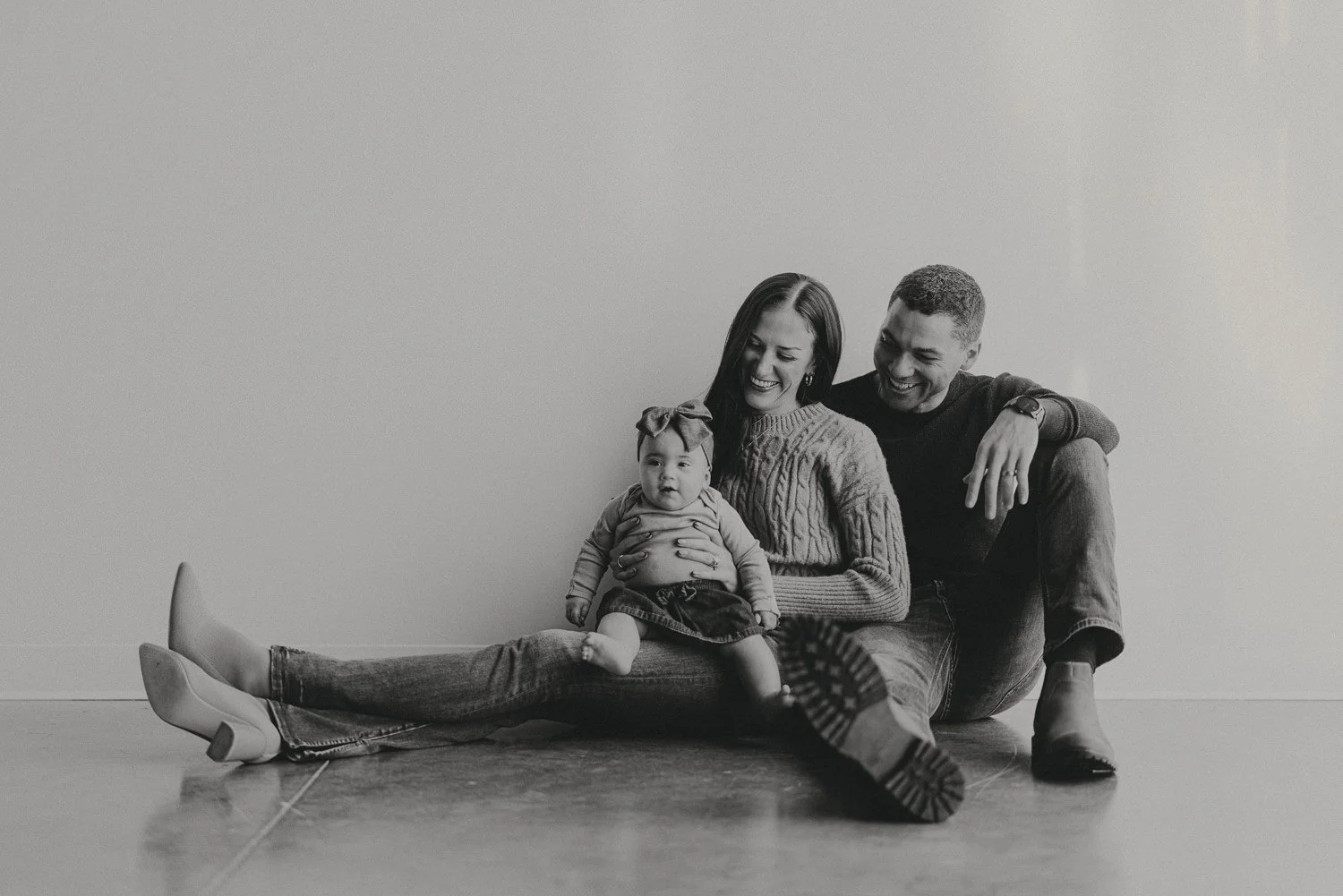 A family of three, consisting of a woman, a man, and a young child, sitting on the floor against a plain wall, smiling and enjoying each other's company. The woman is holding the child on her lap, and all three are casually dressed.