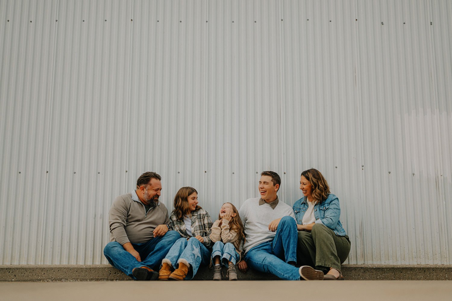 A family of six sitting on the ground in front of a large metal wall, smiling and laughing together.