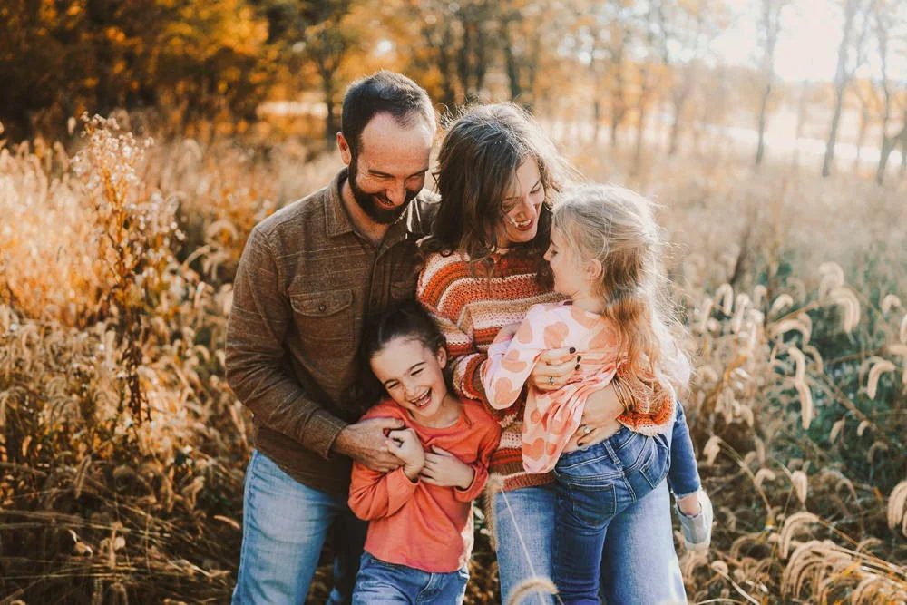 A family of four enjoying a fall day outdoors, hugging and laughing in a field of tall grasses with trees in the background.