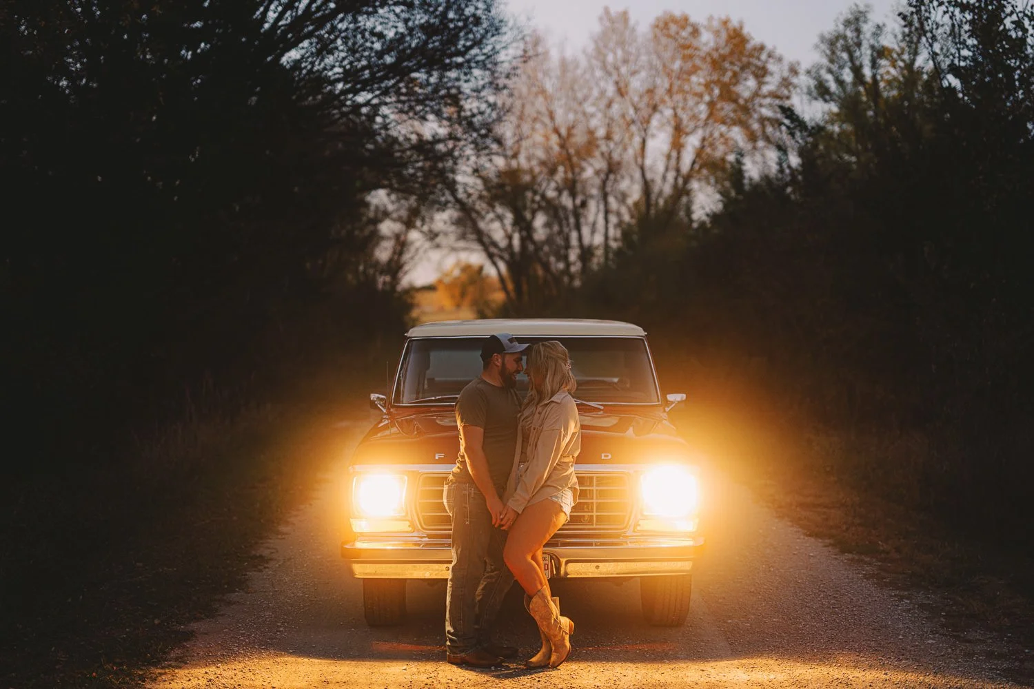 A couple stands close together facing each other in front of a vintage Ford vehicle on a dirt road during sunset, with the vehicle's headlights illuminating them and trees in the background.