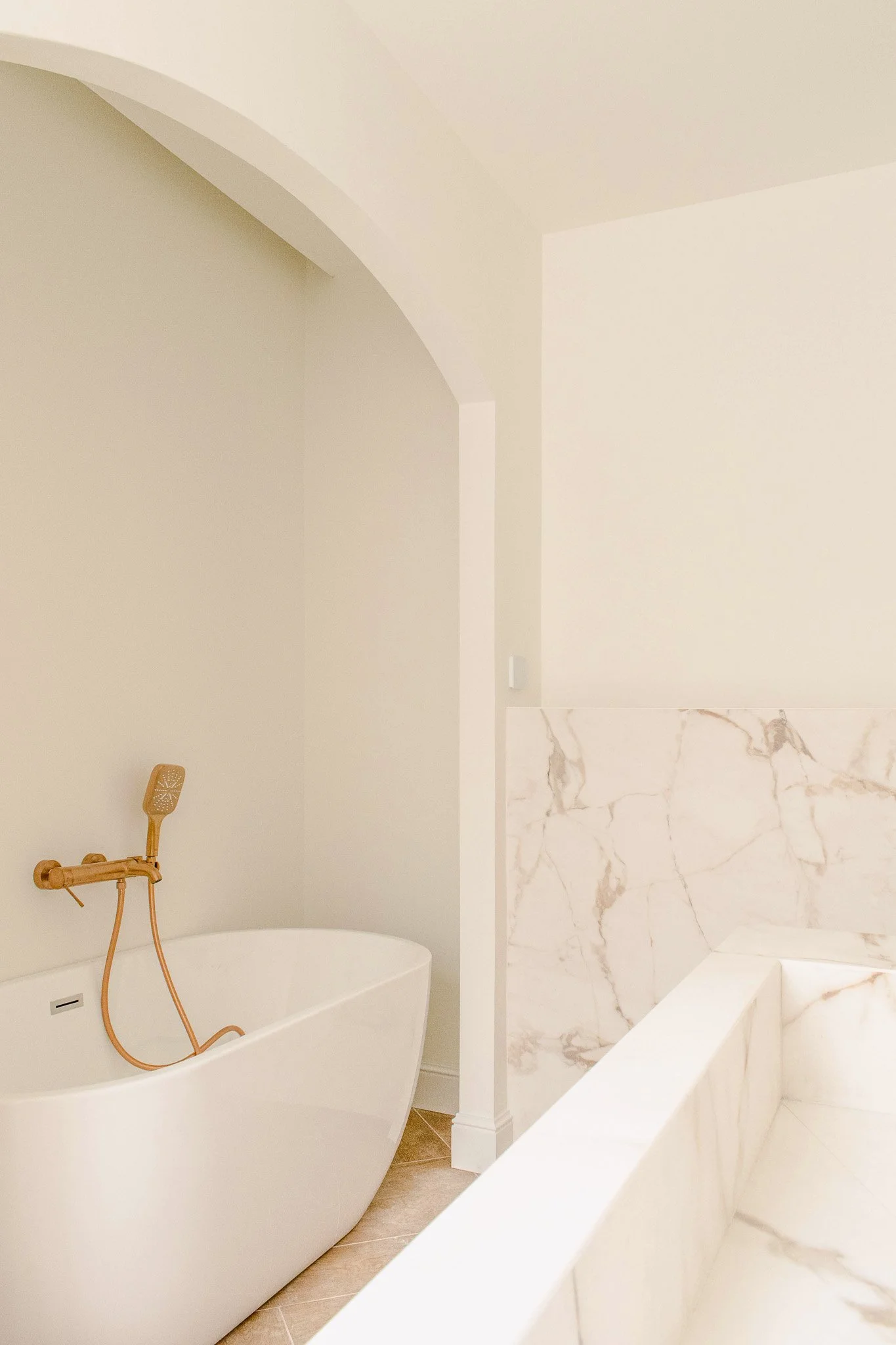 Modern bedroom with a large bed, white bedding, and multiple pillows. The room features a padded headboard with integrated lighting, a small floating nightstand, and a window with a view of palm trees. An arched doorway leads to a bright, minimalistic space with a wall-mounted TV and small sink.