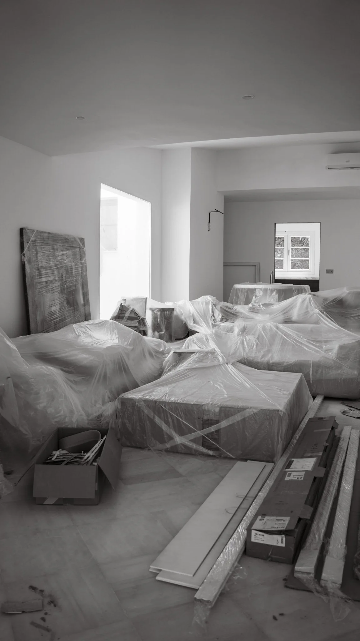 Modern bedroom with a large bed, white bedding, and multiple pillows. The room features a padded headboard with integrated lighting, a small floating nightstand, and a window with a view of palm trees. An arched doorway leads to a bright, minimalistic space with a wall-mounted TV and small sink.