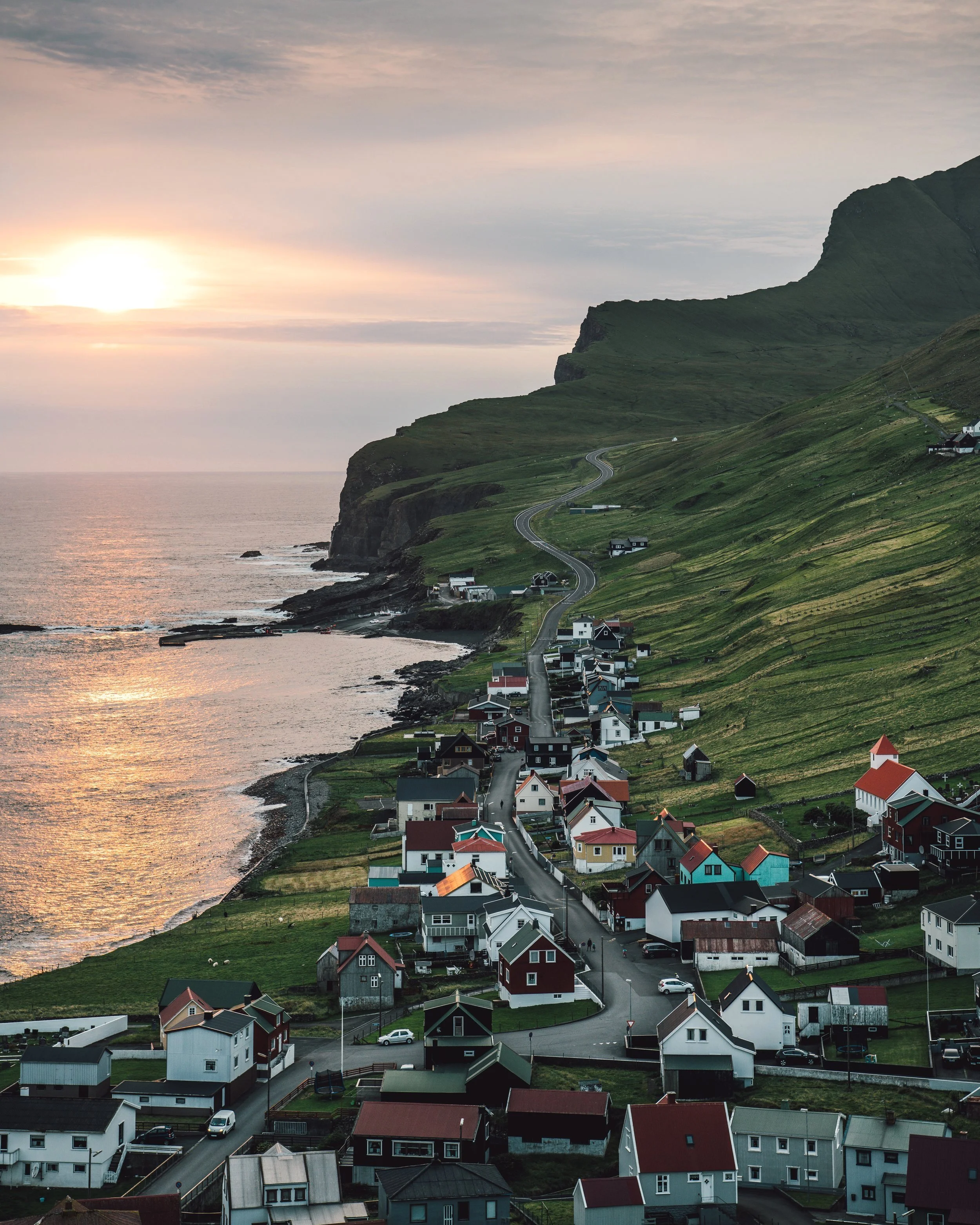 Lille Færøsk by med klassisk færøsk arkitektur, grøn natur og bjerglandskab. Gul solnedgang i baggrunden over havet. Groossartig faglig firmarejse