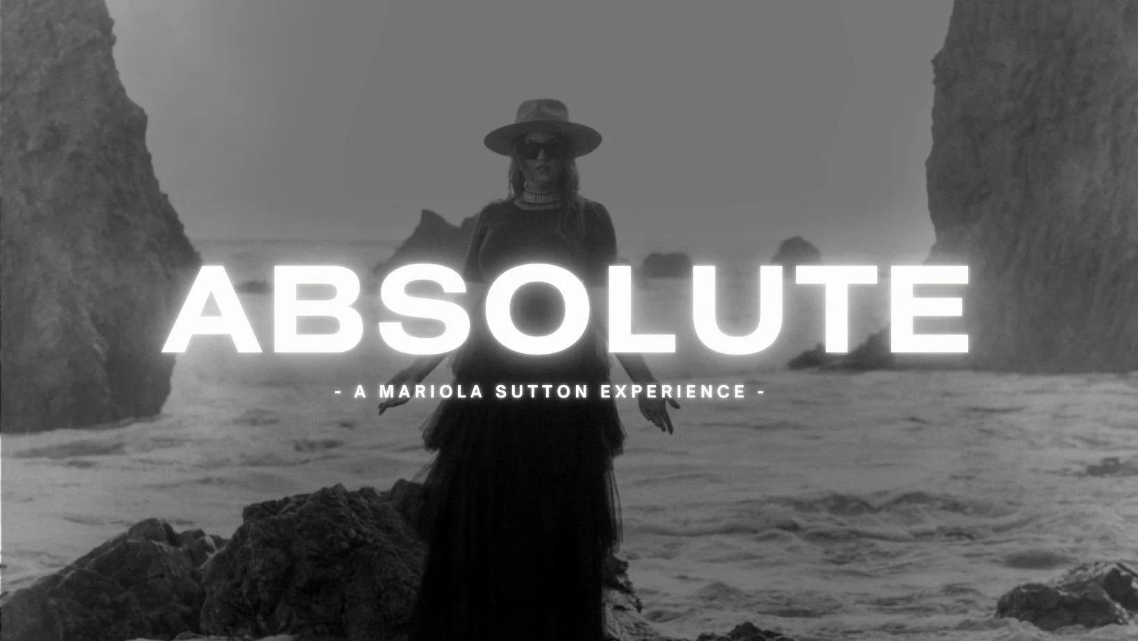 Black and white photo of a woman standing on a rocky beach between two large rock formations, wearing a wide-brimmed hat and sunglasses. The ocean and more rocks are in the background. Large text overlays the image, reading 'ABSOLUTE,' with smaller text underneath that says 'A Mariola Sutton Experience.'