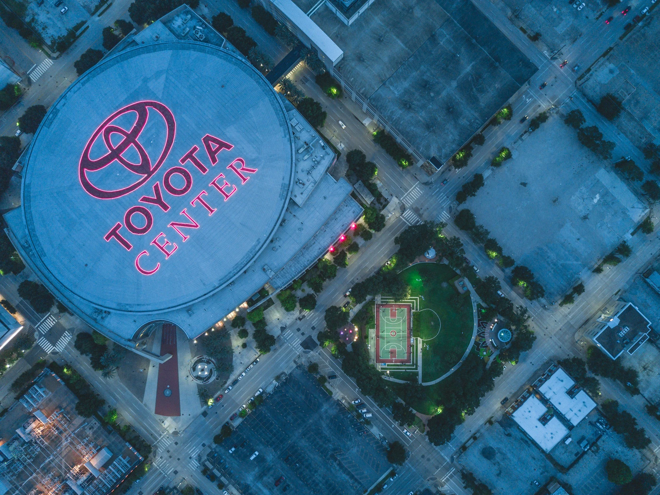 Aerial nighttime view of a city with a large building displaying a neon pink Toyota Center sign, surrounding streets, parking lots, a basketball court, and smaller buildings.