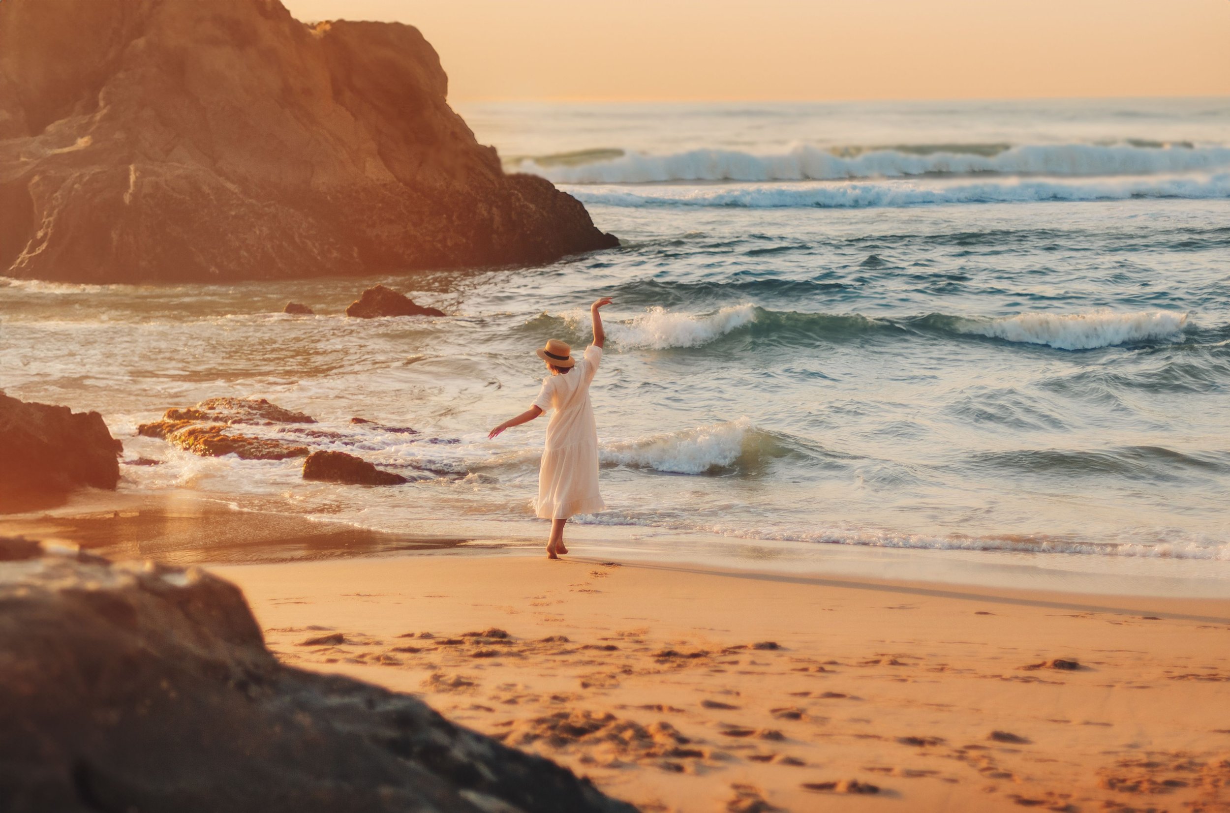Woman in a white dress and a straw hat walking along the beach with waves and a rocky cliff in the background during sunset.