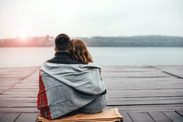 Couple sitting on a dock with a blanket, overlooking a lake at sunset.