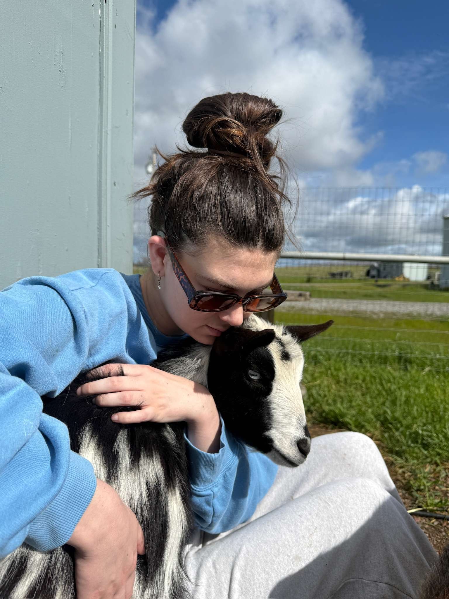 A woman in sunglasses hugs a baby goat outdoors against a green fence and blue sky with clouds.