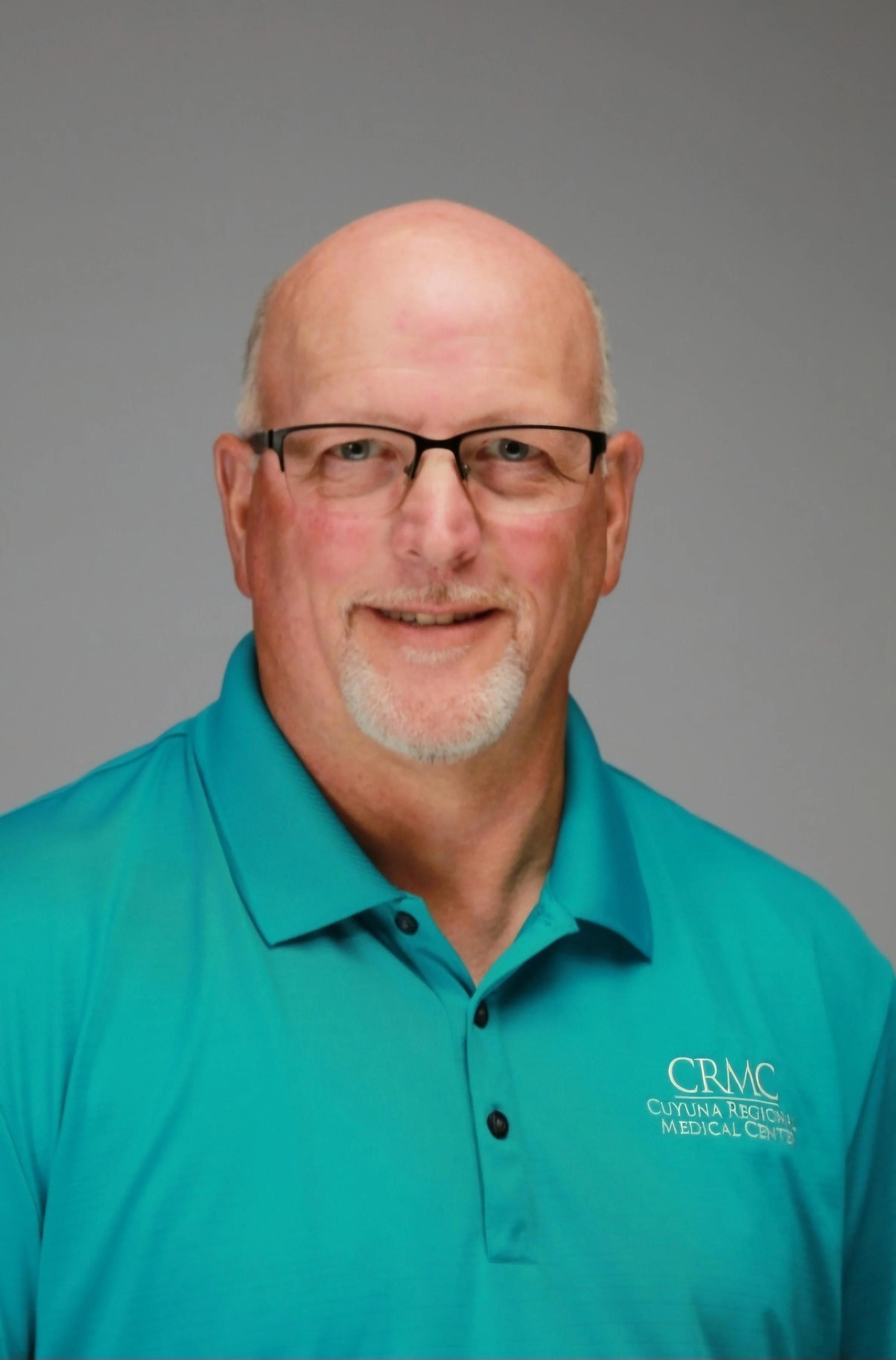Close-up of a bald man with glasses, smiling, wearing a teal polo shirt with the logo 'CRMC Cuyuna Regional Medical Center' on the chest, against a plain gray background.
