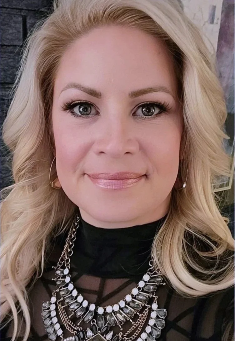 Close-up of a woman with blonde hair, wearing makeup, earrings, a black top, and layered necklaces, smiling at the camera.