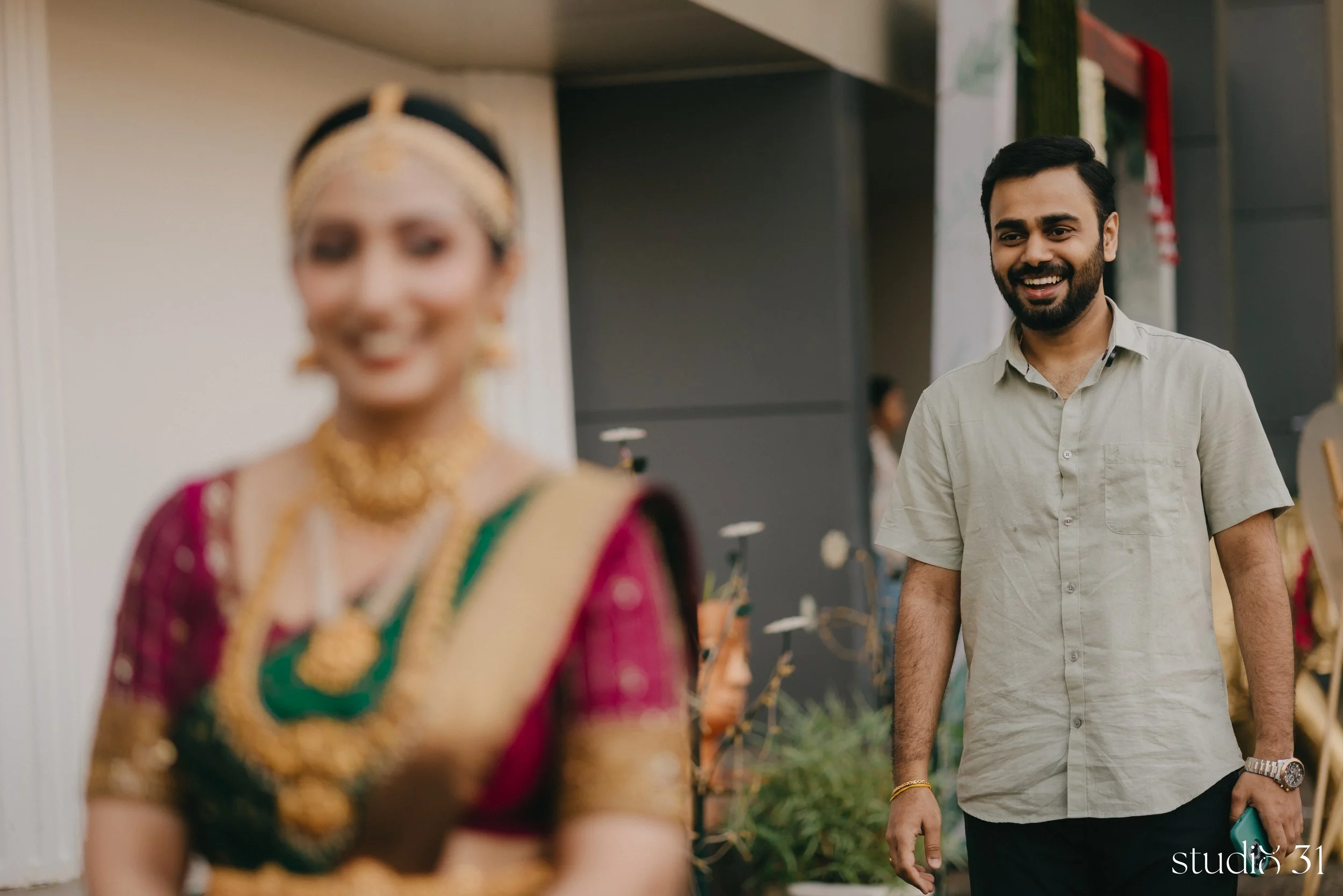 A man with a beard and dark hair smiling while looking at a woman in traditional Indian attire. The woman's face is blurred, and she is wearing a colorful sari with elaborate jewelry. They appear to be at an event or celebration.