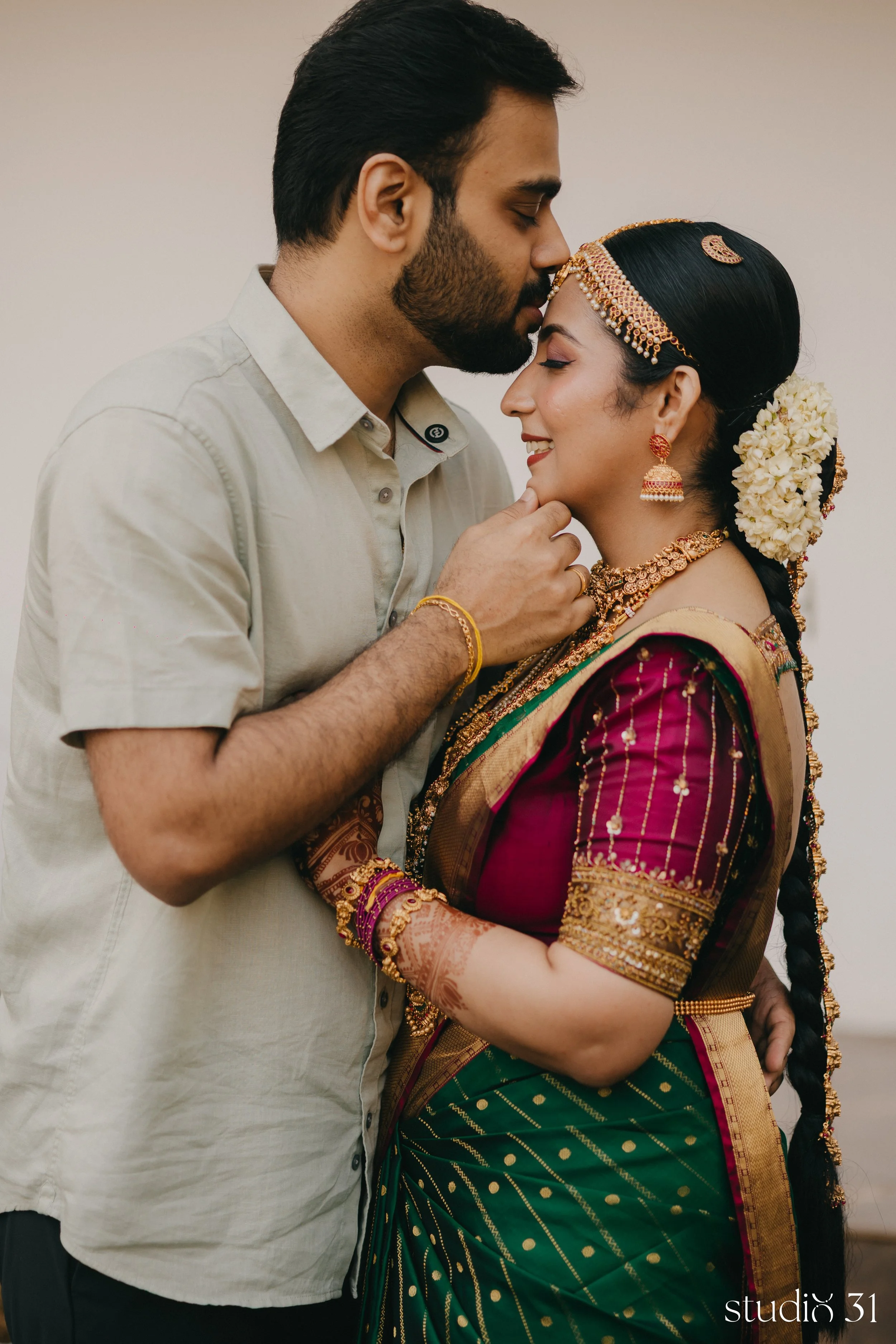 A man and woman are sharing an intimate moment, with the man gently kissing the woman's forehead. The woman is dressed in traditional Indian attire, adorned with intricate jewelry and flowers in her hair, while the man is wearing a light-colored shirt. They both have their eyes closed and appear to be very happy.