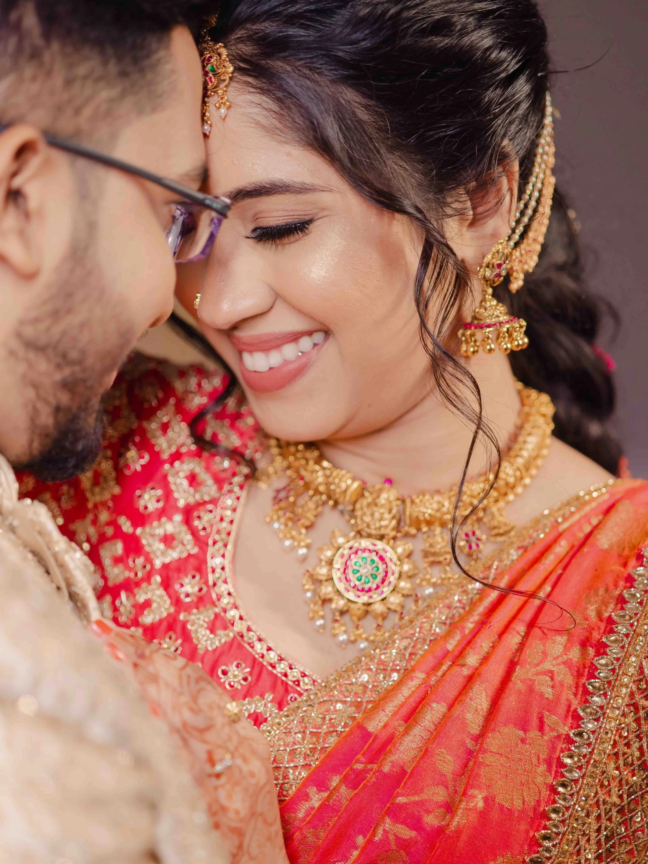A close-up of a smiling woman dressed in traditional Indian attire with gold jewelry, including earrings, necklace, and headpieces, during a celebration or wedding.