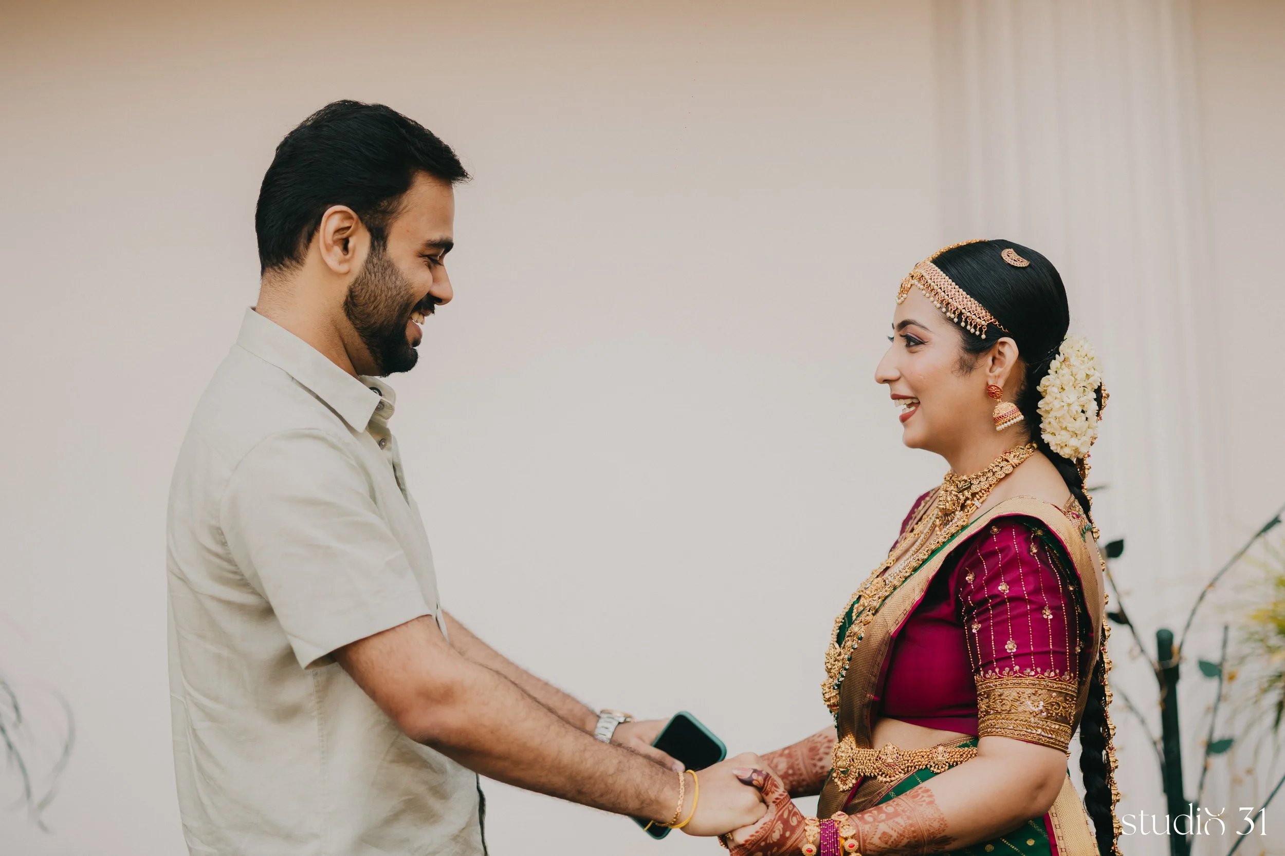 A man and woman standing face to face, holding hands, during a wedding ceremony. The woman is dressed in traditional Indian attire with gold jewelry and flowers in her hair, and the man is casually dressed in a light-colored shirt.