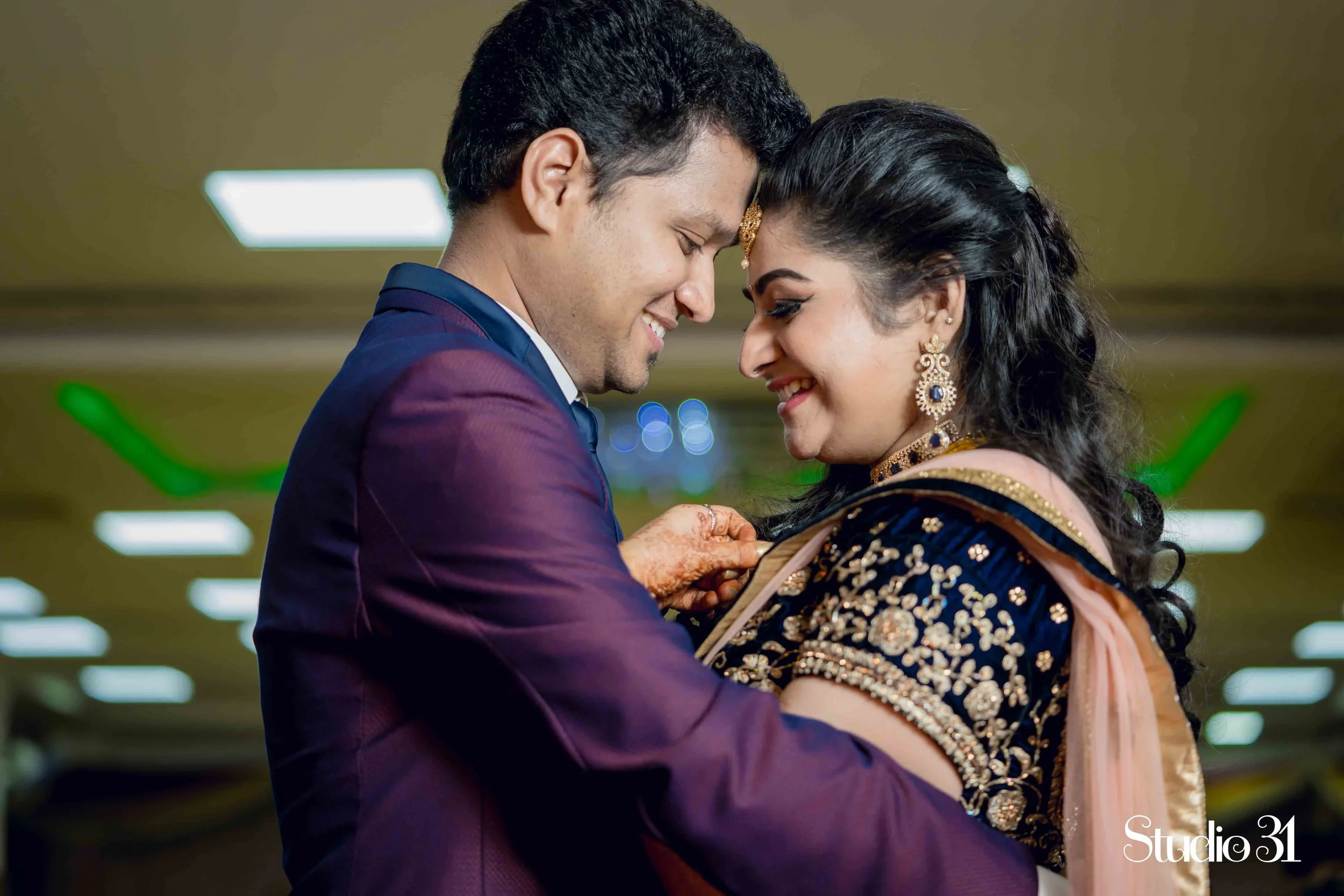 A couple dressed in traditional Indian attire smiling and touching foreheads in a dance pose inside a building with fluorescent lights.