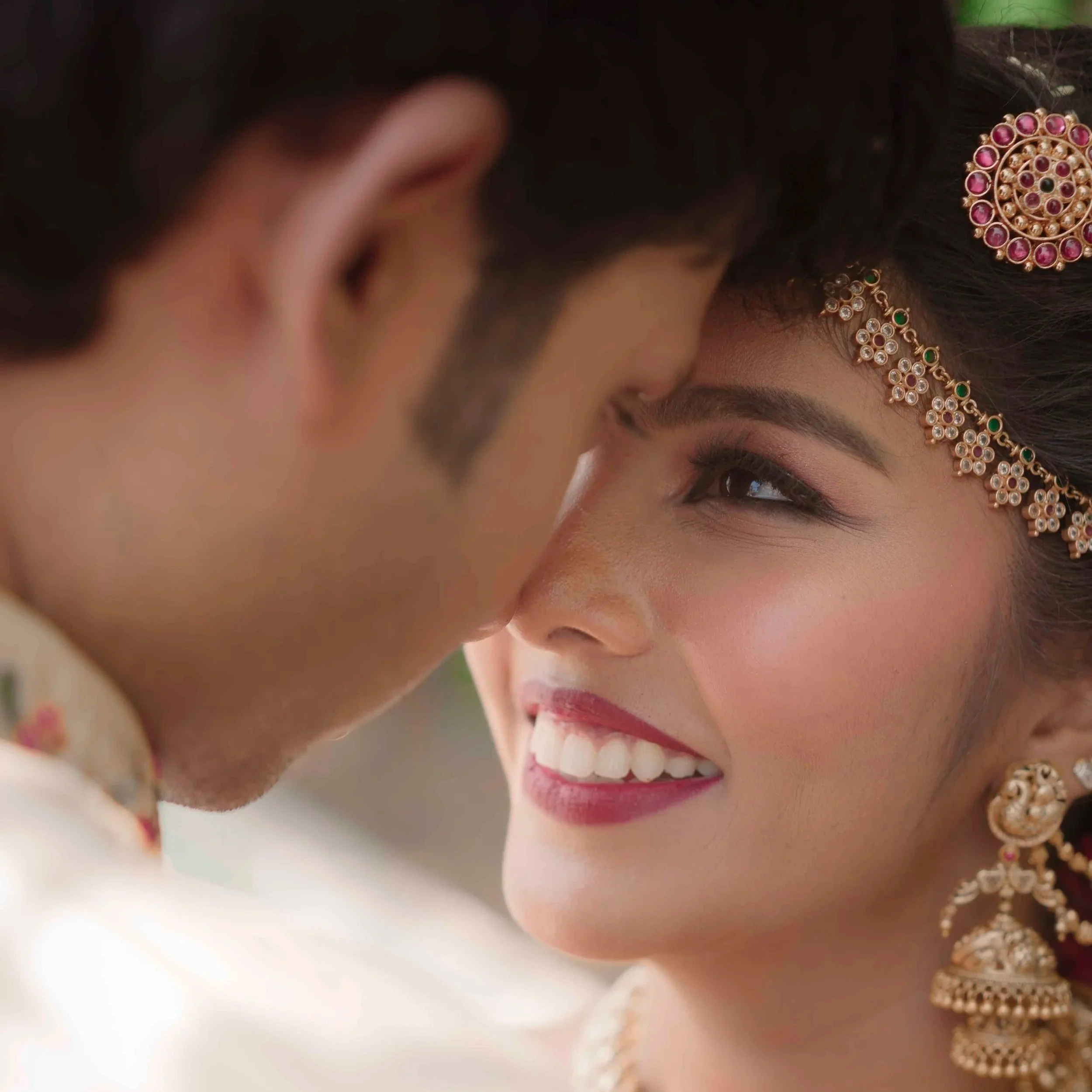 Close-up of a smiling woman wearing traditional jewelry, including earrings and a forehead ornament, with a blurred background.