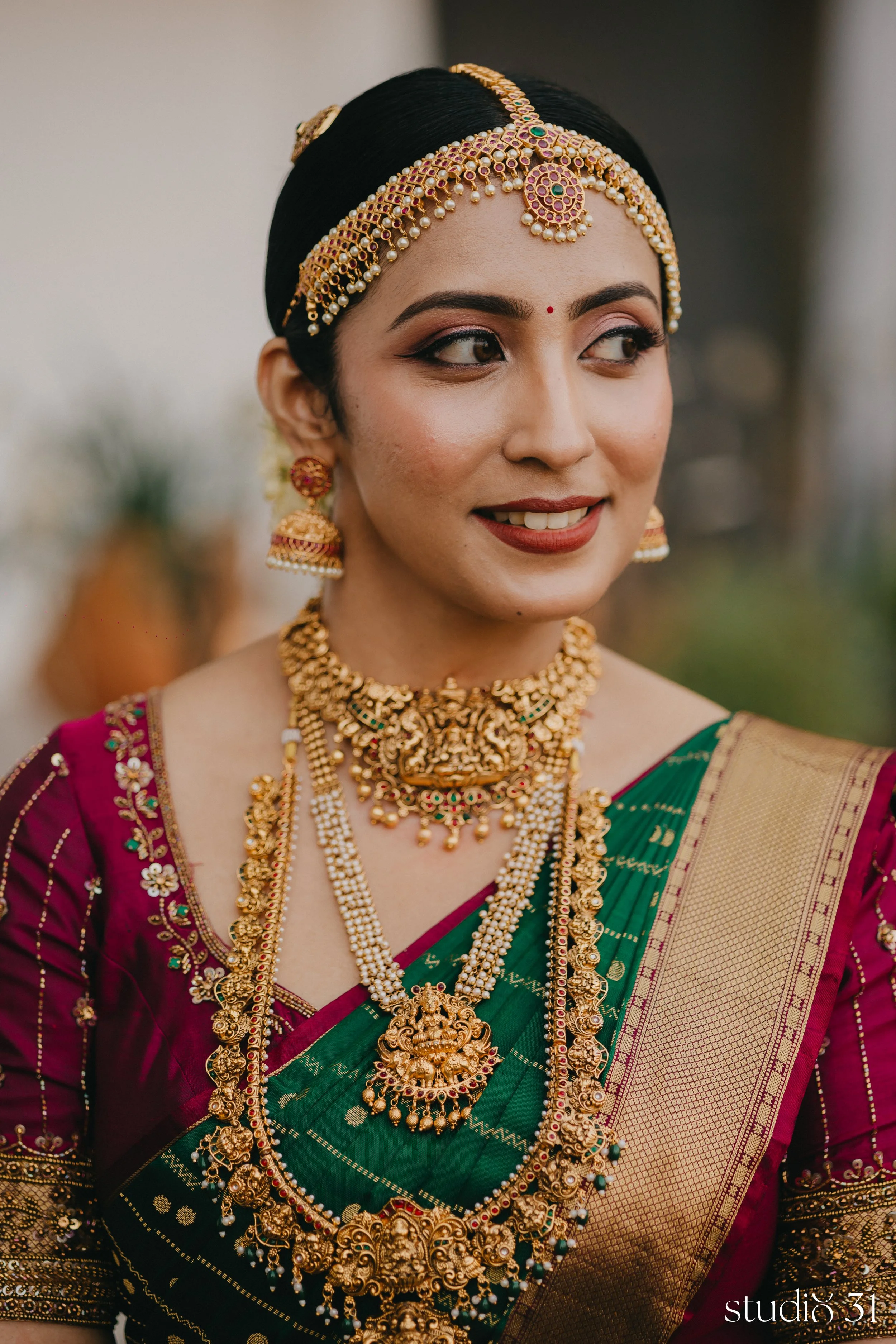 Close-up of a woman wearing traditional Indian saree and elaborate gold jewelry, including necklaces, earrings, and a headpiece, smiling slightly.