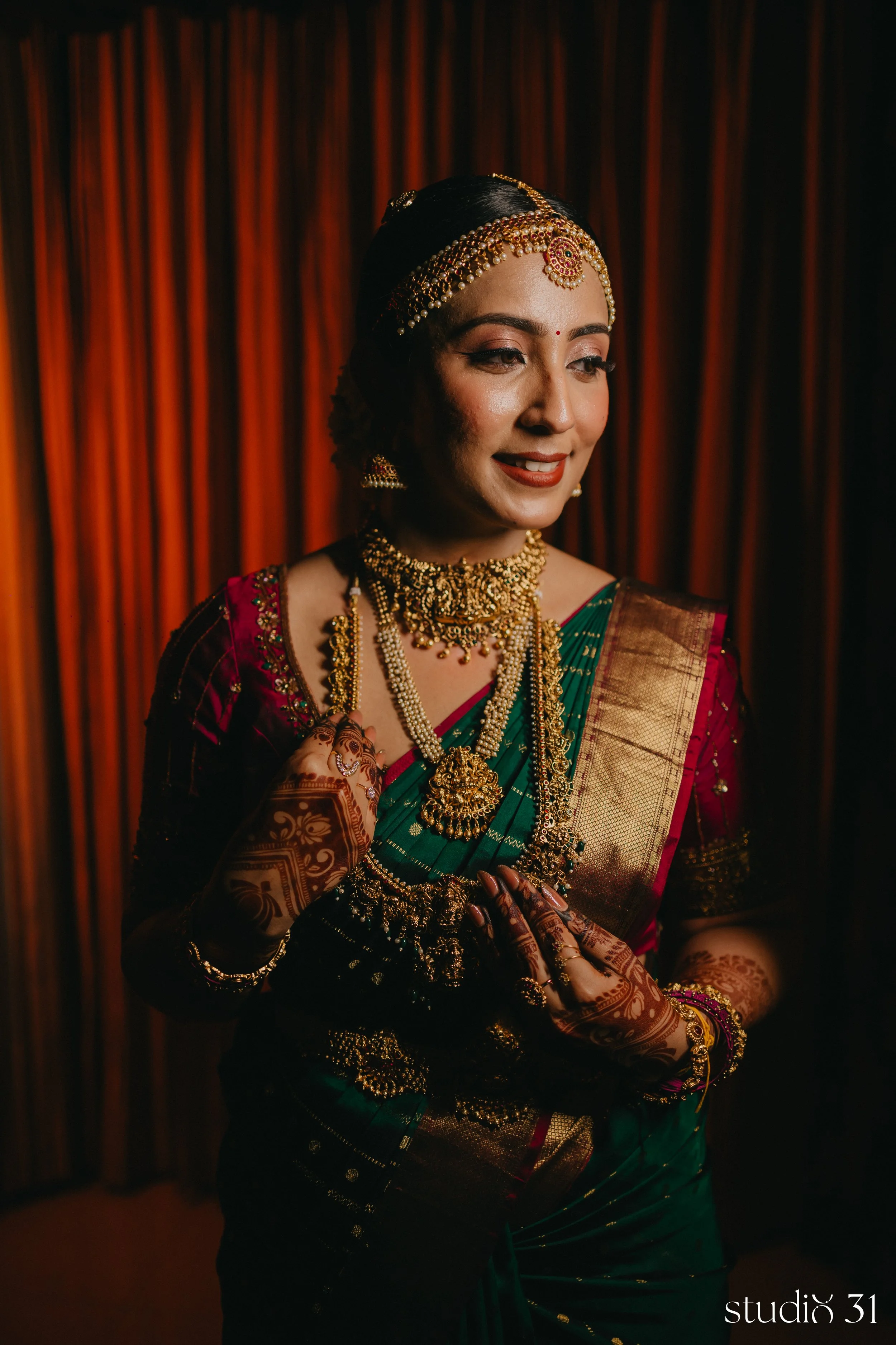 Close-up of a woman dressed in traditional Indian attire with intricate jewelry, mehndi on her hands, and a dark background with red curtains.