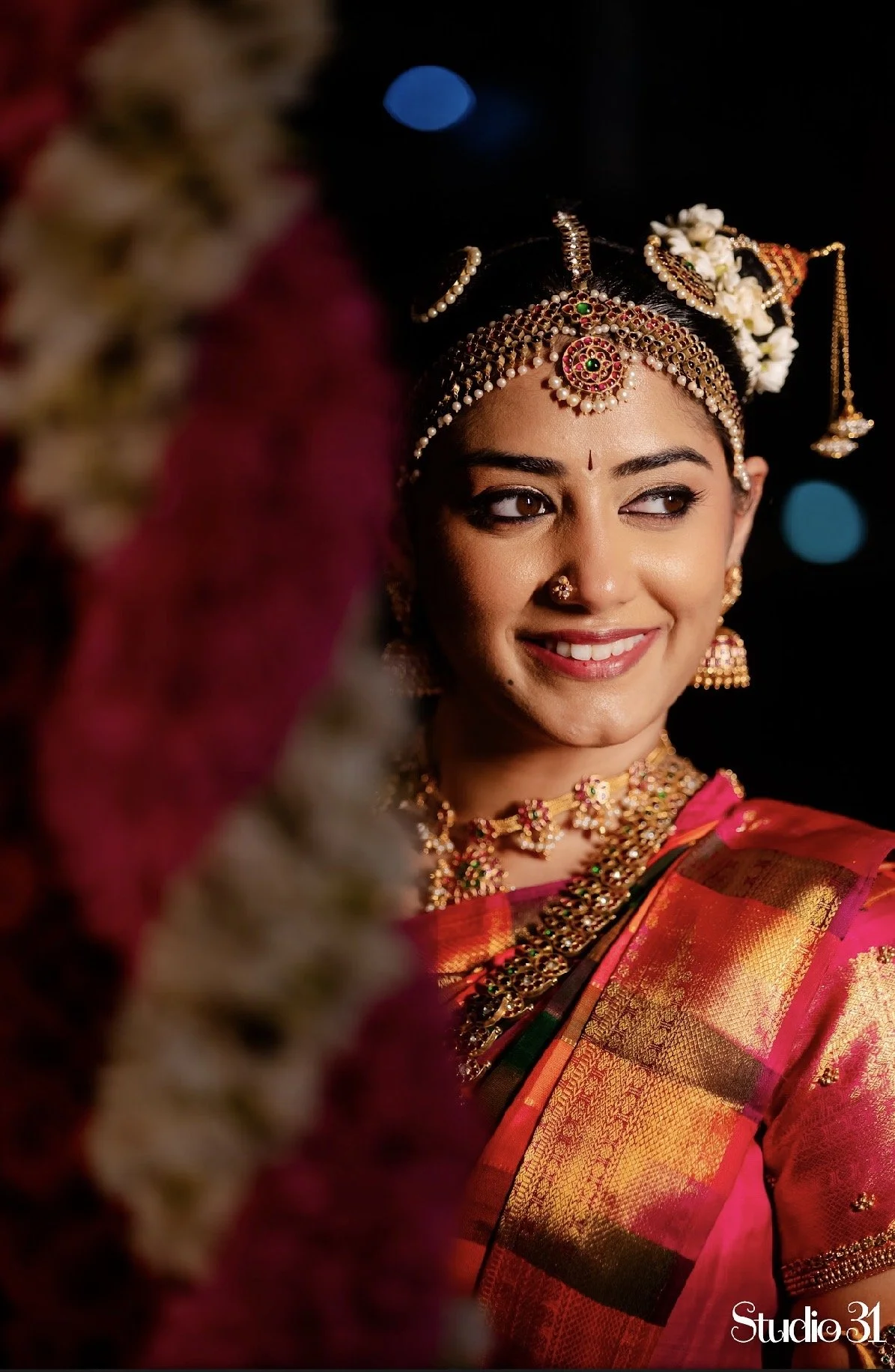 A woman dressed in traditional Indian attire wearing intricate gold jewelry, a vibrant red and orange silk saree, and floral adornments in her hair, smiling with a happy expression.