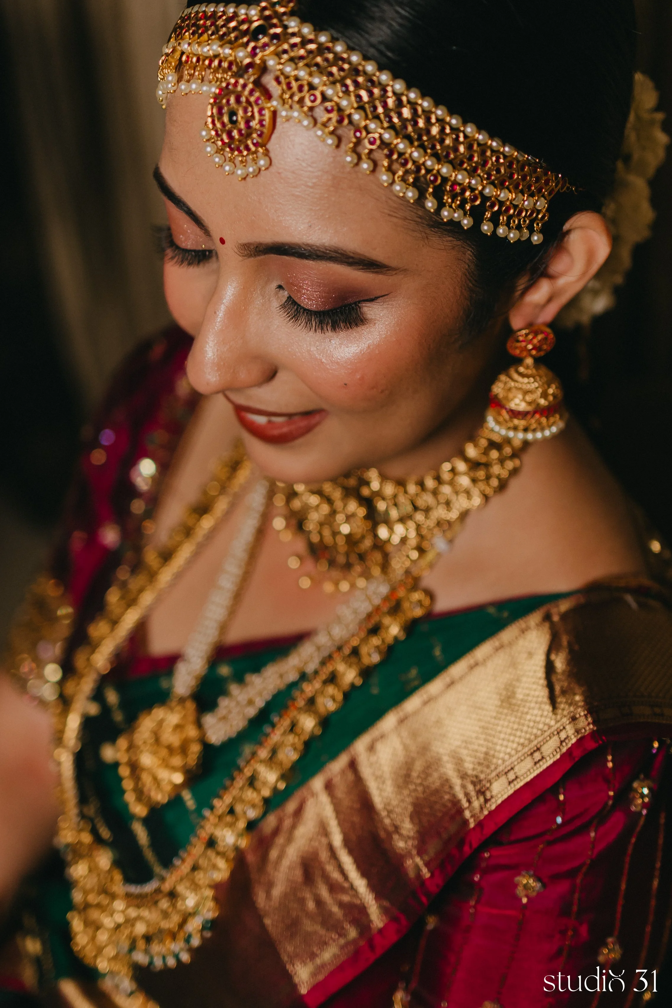 Close-up of a woman's face and upper body dressed in traditional Indian bridal attire with elaborate jewelry and makeup, with closed eyes and a gentle smile.