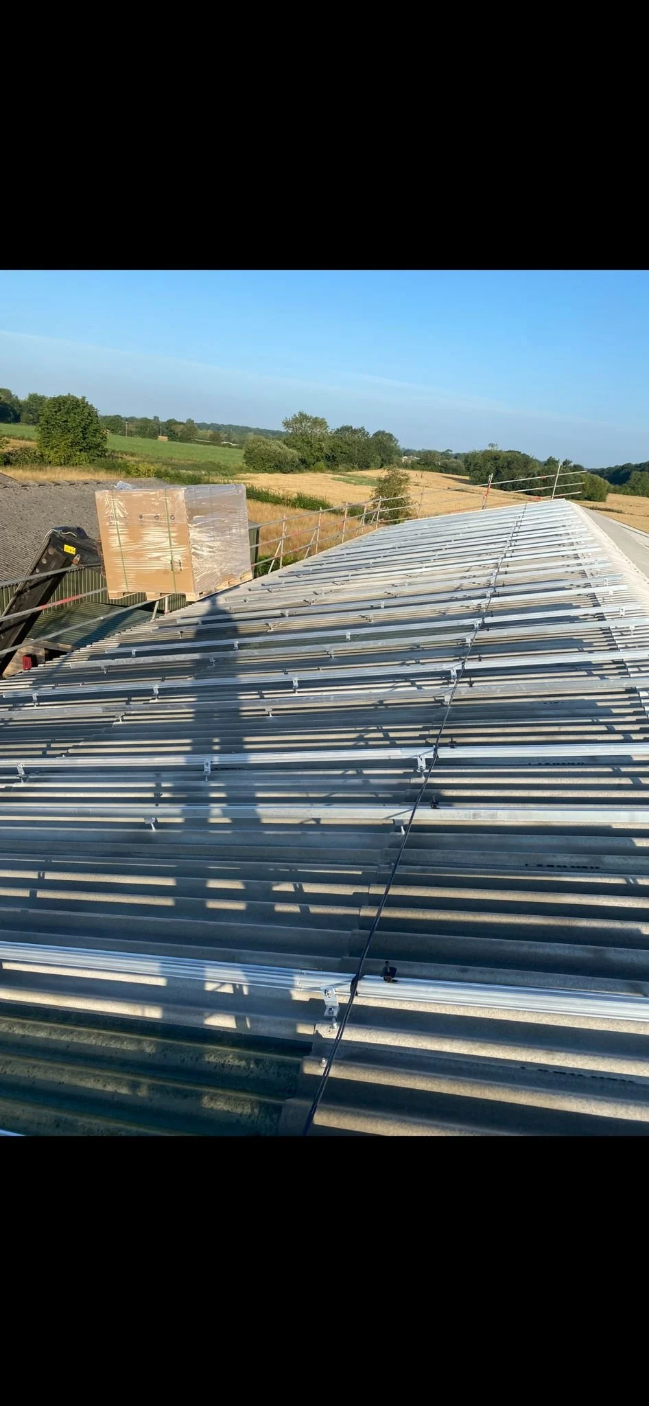 Construction site with a curved metal roof under sunlight, casting shadows, with fields and trees in the background.