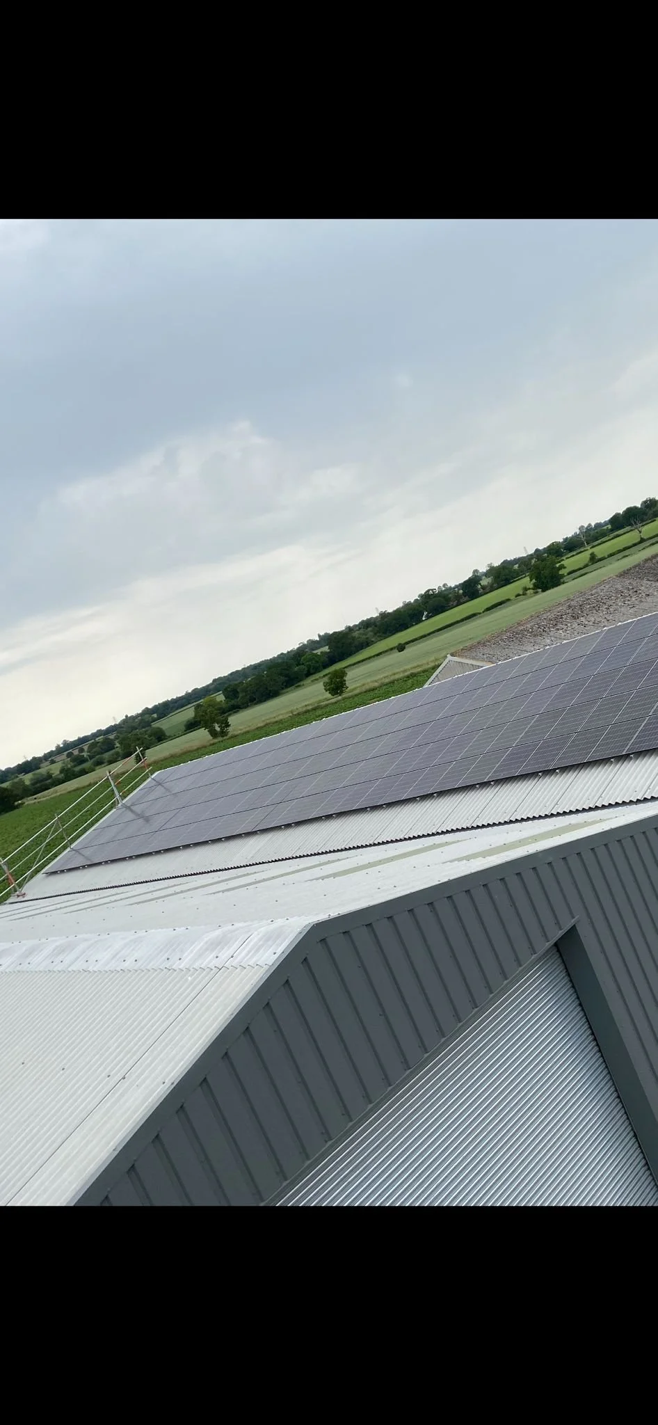 Photograph of a building with solar panels on the roof, set against a rural landscape with trees and open fields under a cloudy sky.