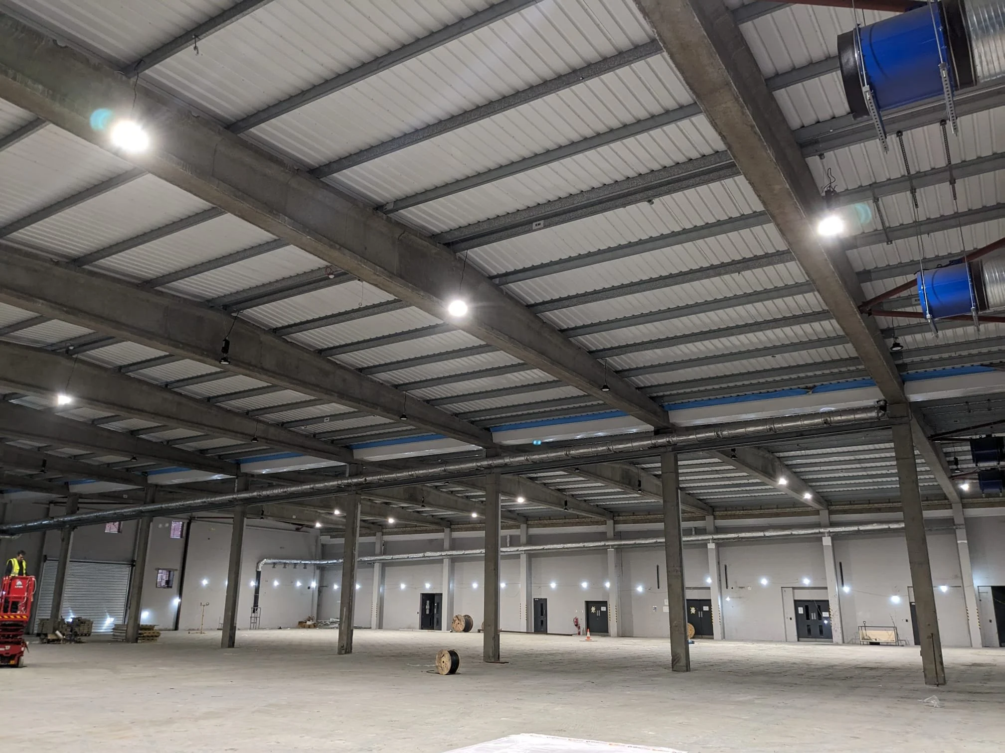 Large empty industrial warehouse with steel beams, bright overhead lights, and a concrete floor, some construction materials and equipment are visible.