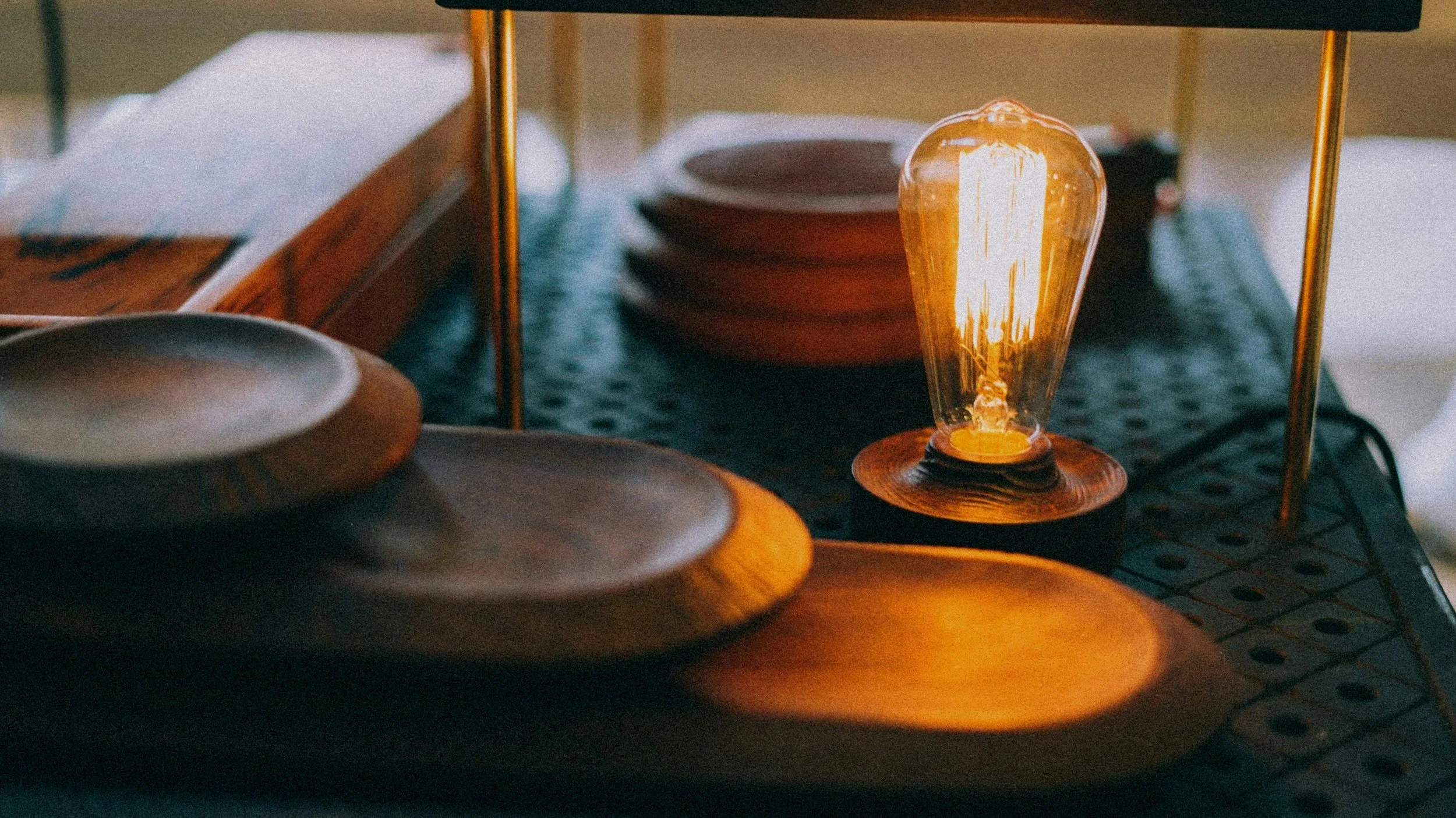 A warm glowing filament light bulb on a dark surface, with a stack of brown bowls in the background and a reflective mirror behind it. The scene has a cozy, vintage aesthetic.