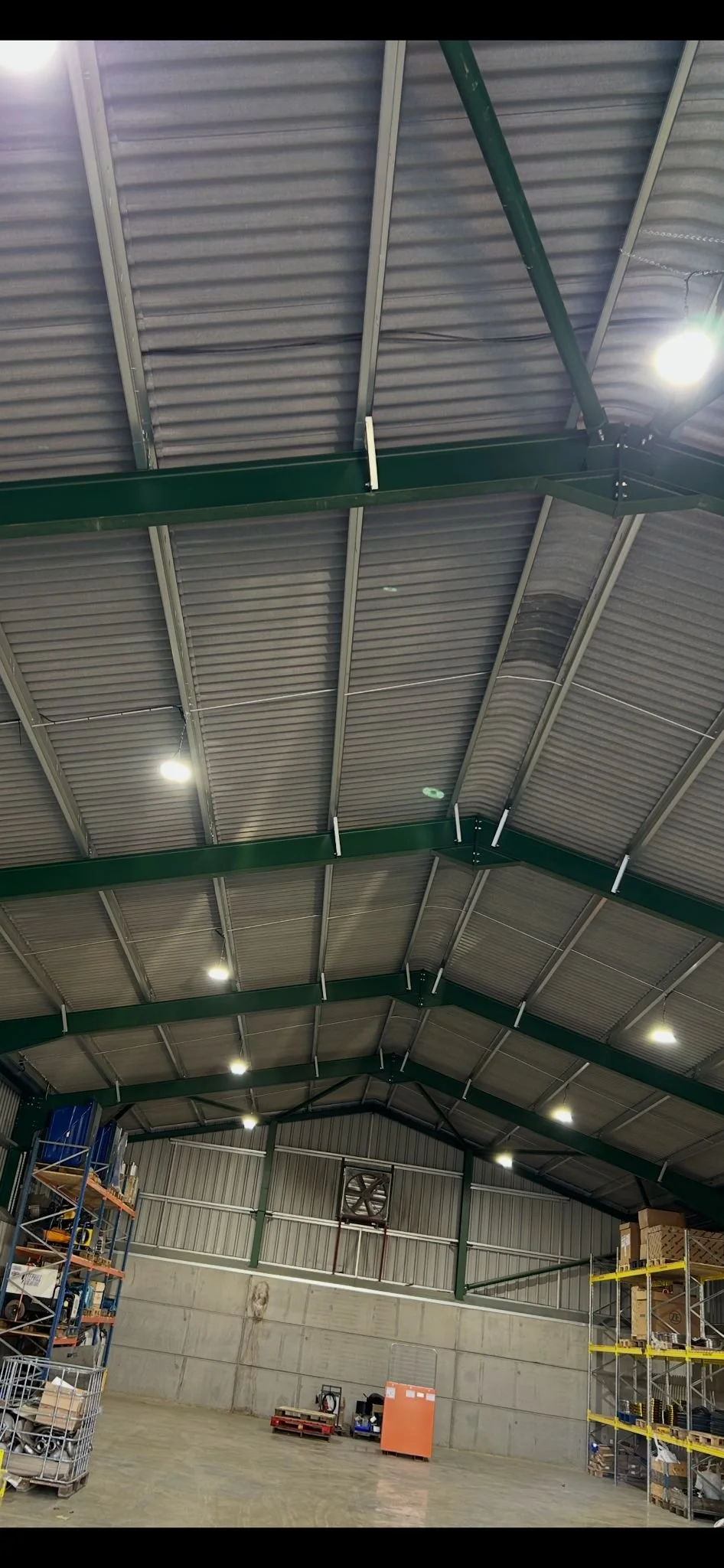 An industrial warehouse interior with metal shelving and storage bins on the left, a fuse box and some pallets in the back, and bright ceiling lights overhead.