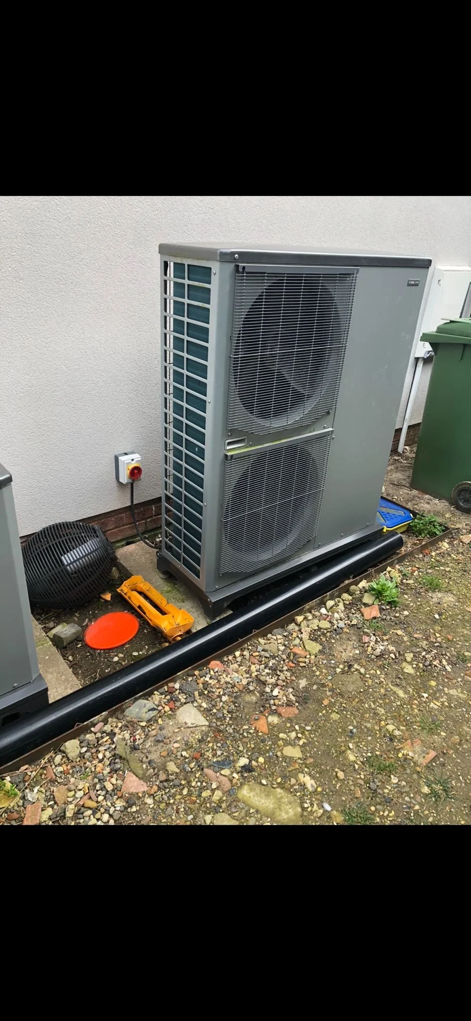 An outdoor air conditioning unit with two large fans on its front side, installed beside a beige exterior wall, with a green trash bin and small tools on the ground nearby.